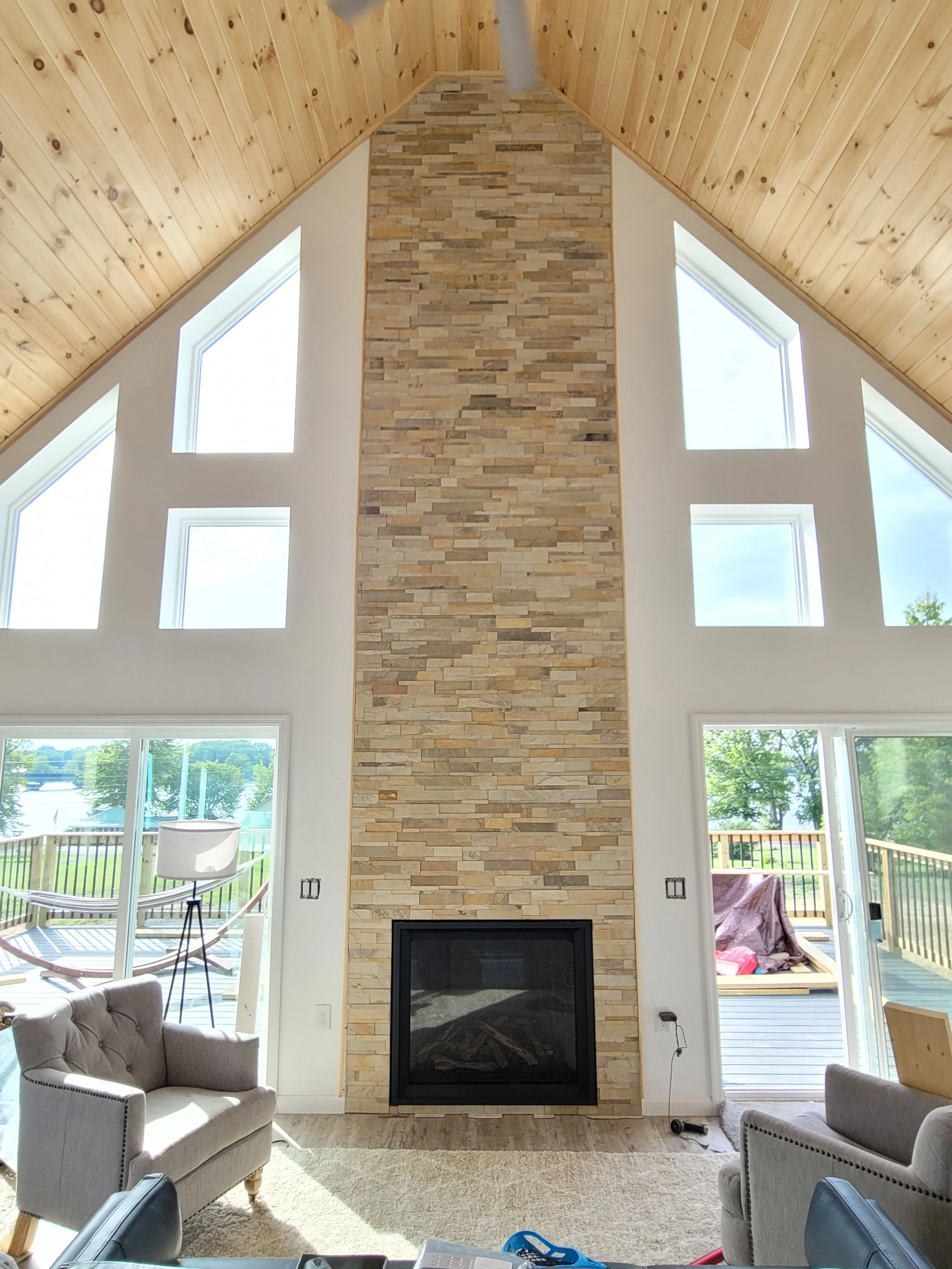 A living room with a high, sloped wooden ceiling, a stone fireplace, and large windows letting in natural light. There are two beige armchairs, a floor lamp, and outdoor patio furniture visible through the windows.