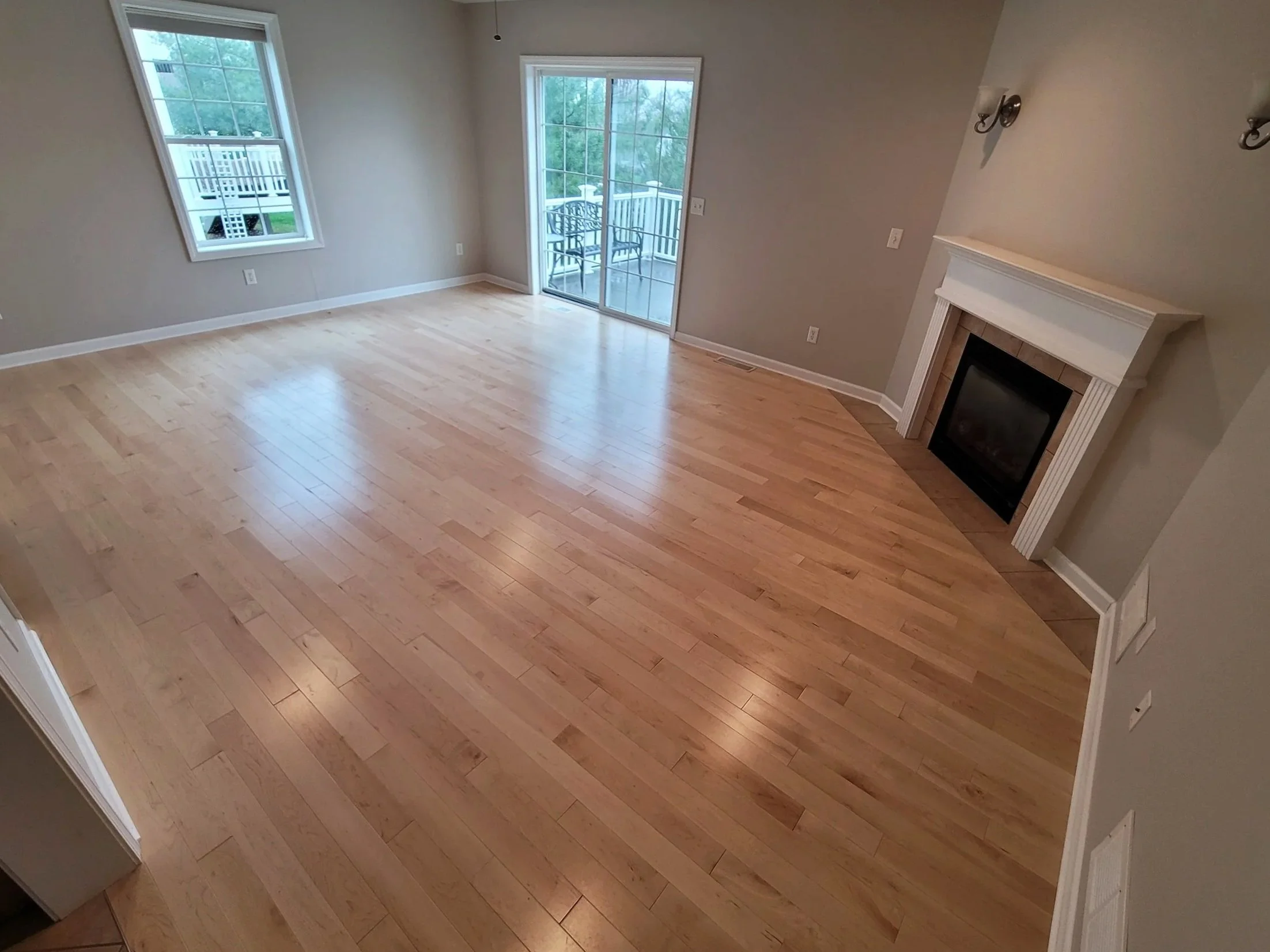 Empty living room with large windows, a sliding glass door leading to a balcony, hardwood floors, a fireplace with a white mantle, and wall-mounted light fixtures.