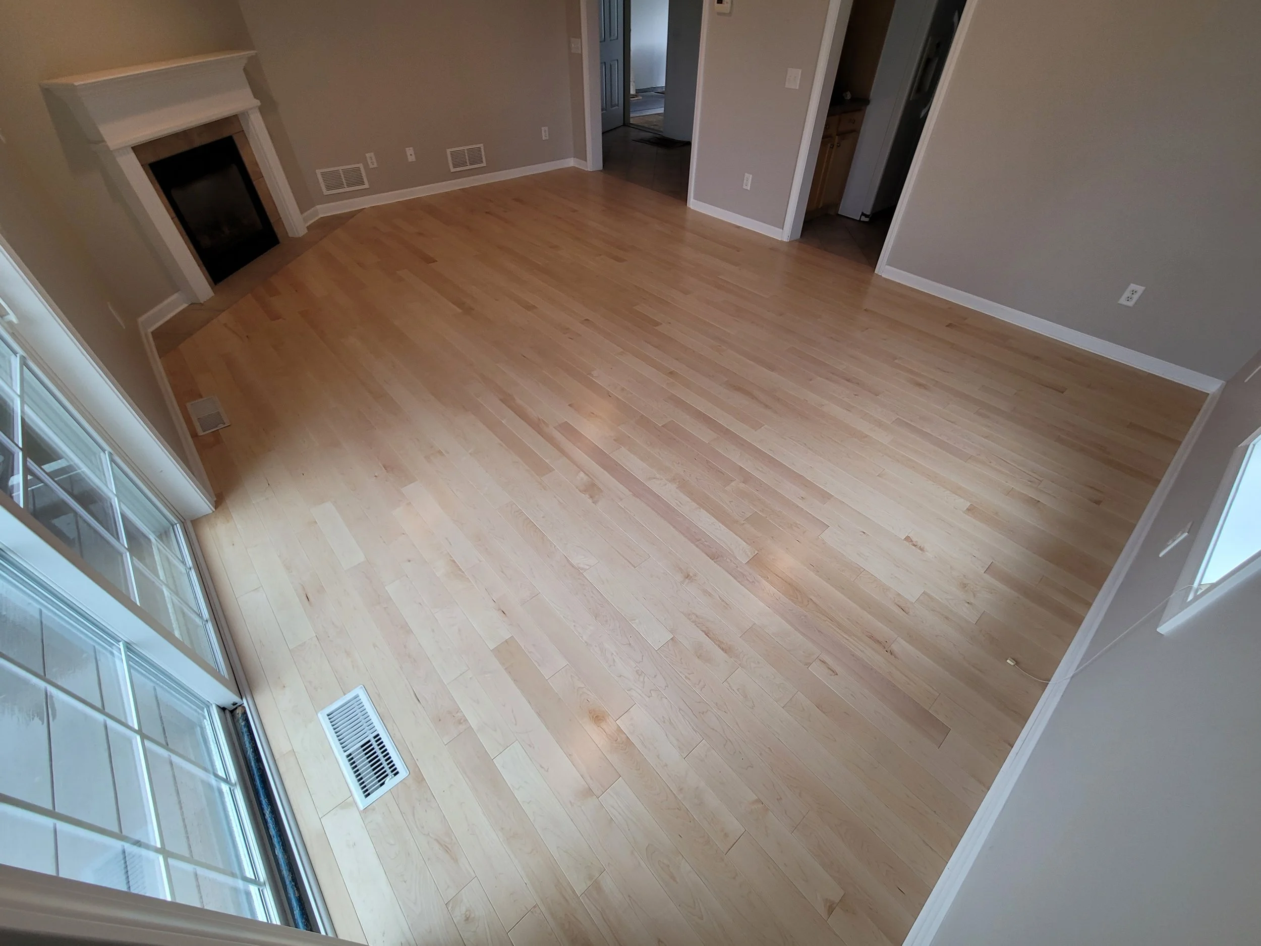 Empty living room with light wooden floor, beige walls, a fireplace, and a large window.