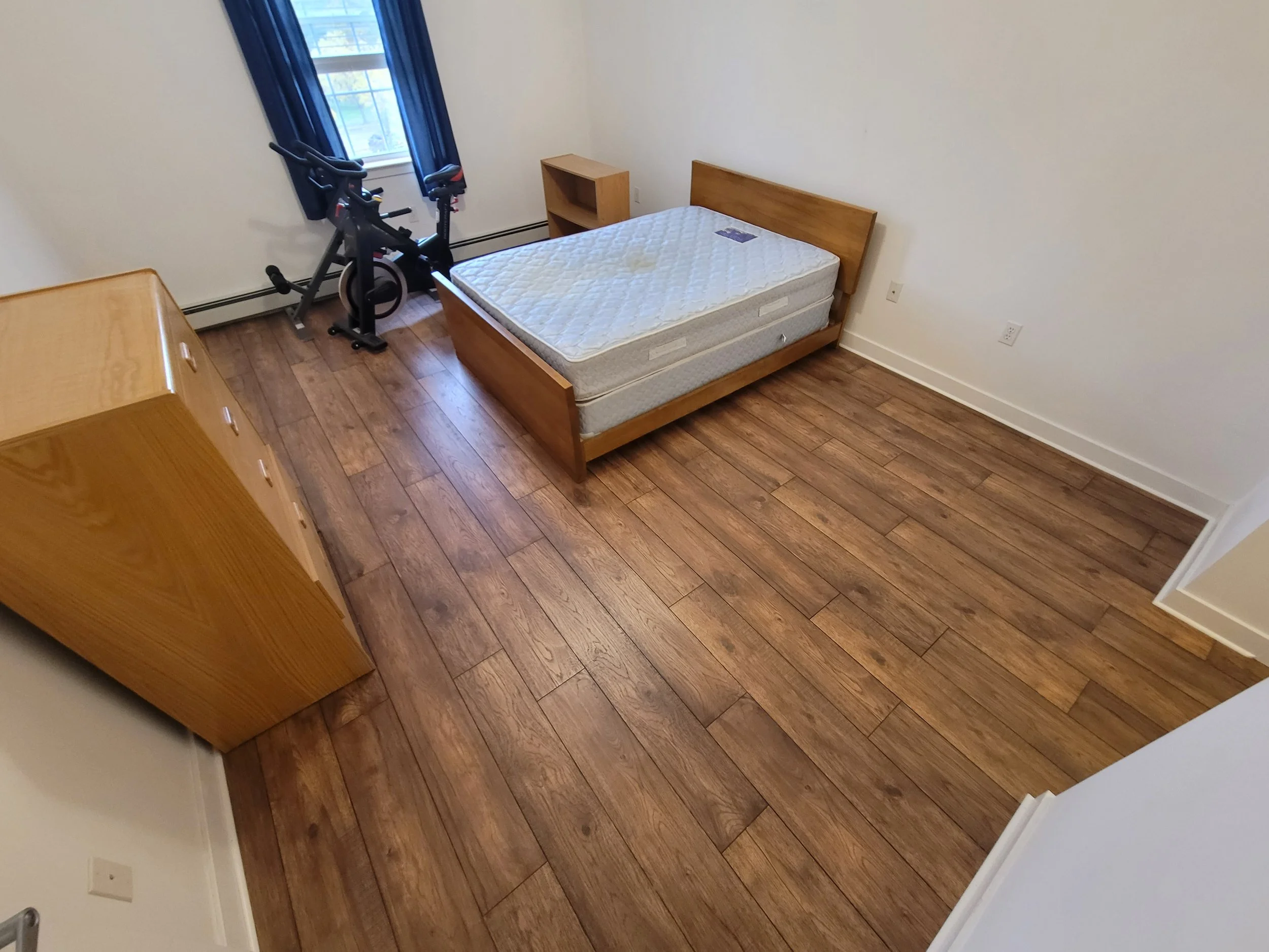 A small bedroom with a wooden bed frame and mattress, a wooden dresser, two workout bikes near the window, wood flooring, and a small wooden bookshelf.