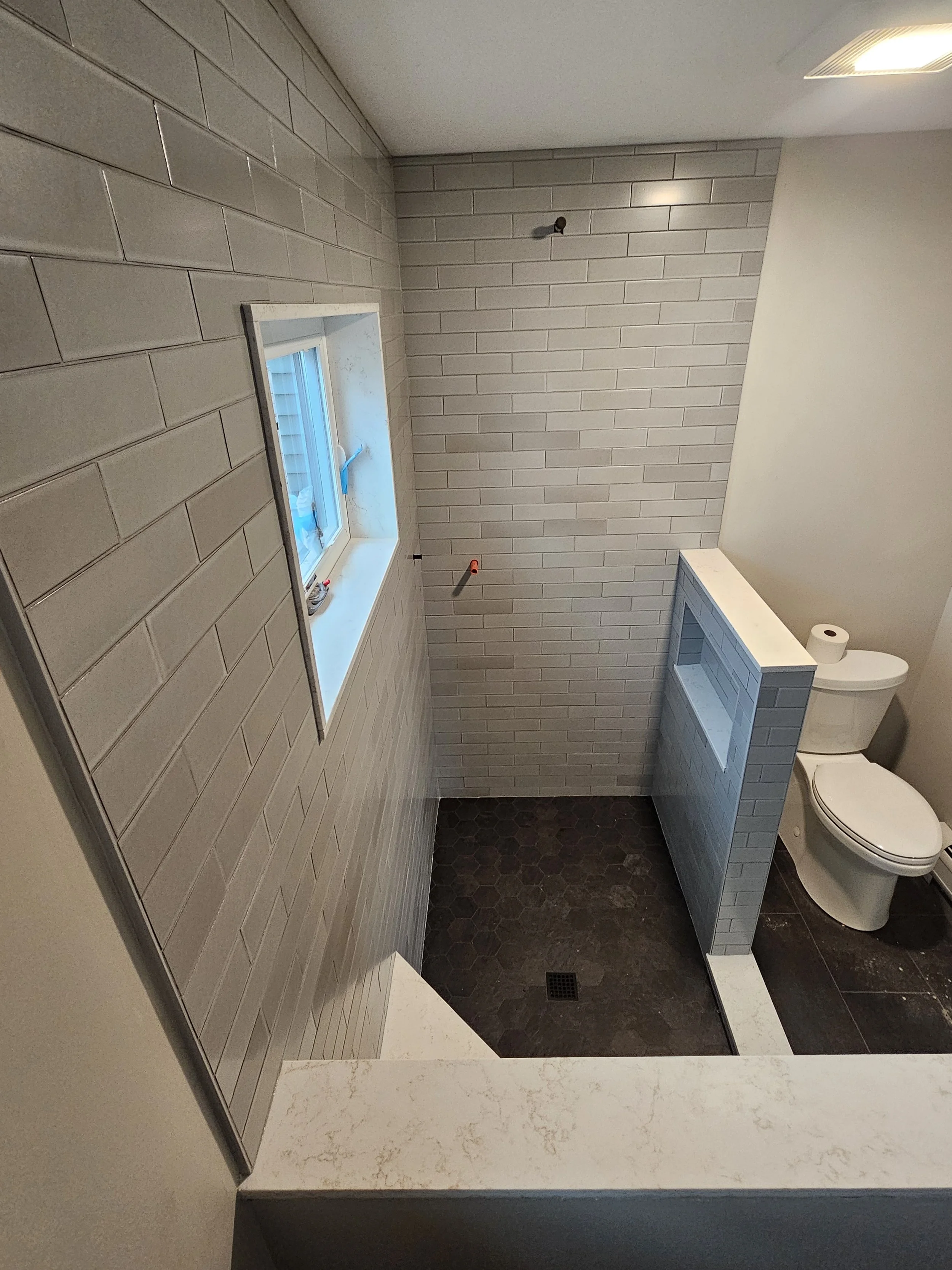 Bathroom under construction with beige subway tile walls, dark hexagonal tile floor, a small window, toilet, and a half-wall with a towel holder.
