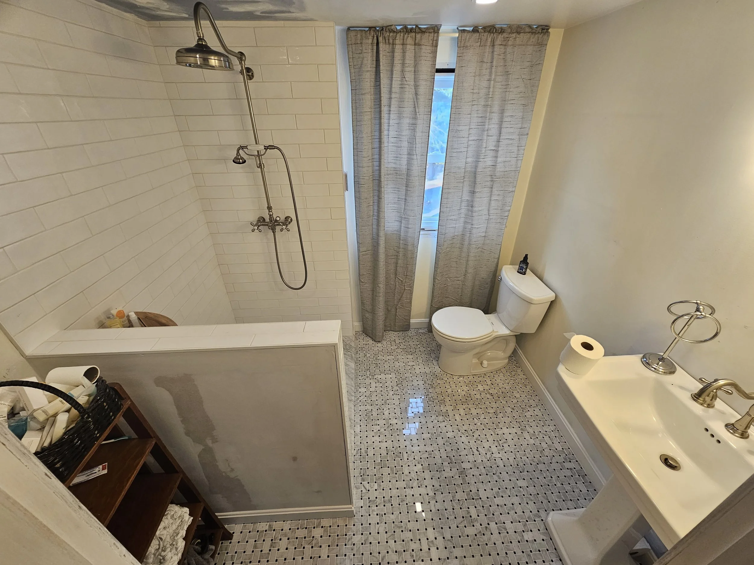 A small bathroom with a walk-in shower, a toilet, a vanity with a sink, a mirror, and window with curtains. There are personal care items in a basket and on the vanity.