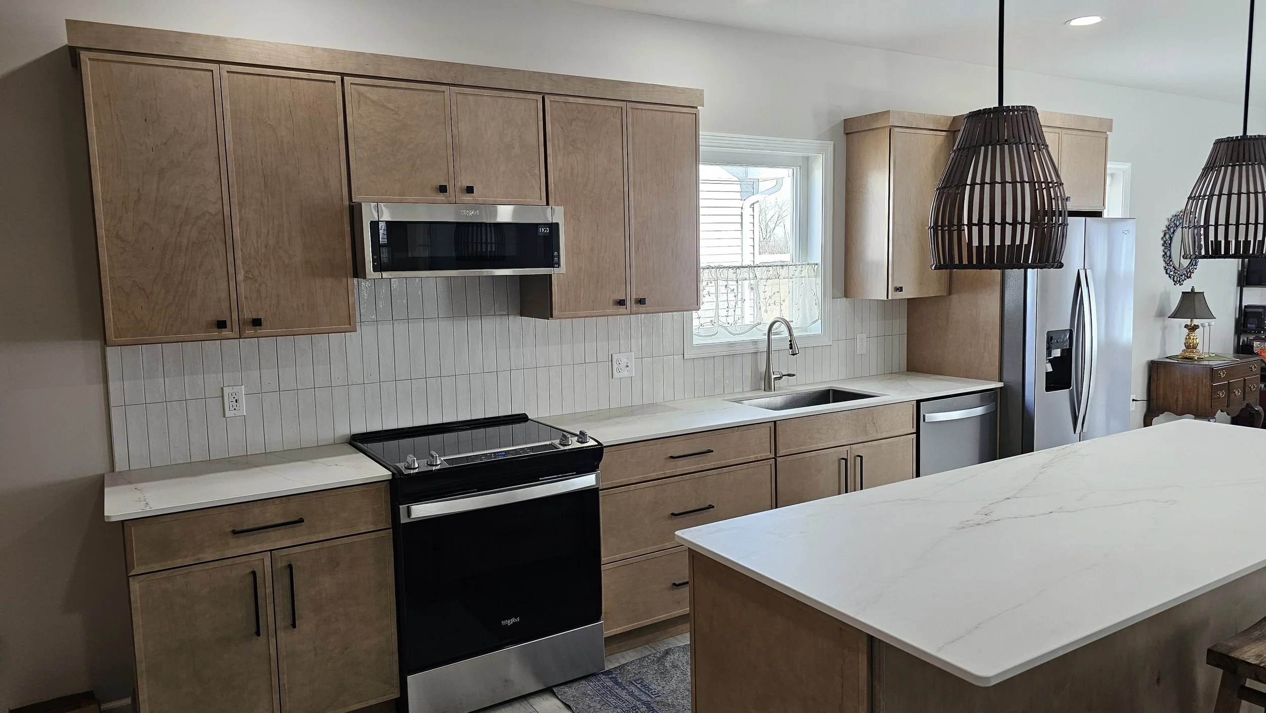 Kitchen with light wood cabinets, a white marble countertop, stainless steel appliances including a refrigerator, oven, and microwave, and a window above the sink. Two large woven pendant lights hang from the ceiling.