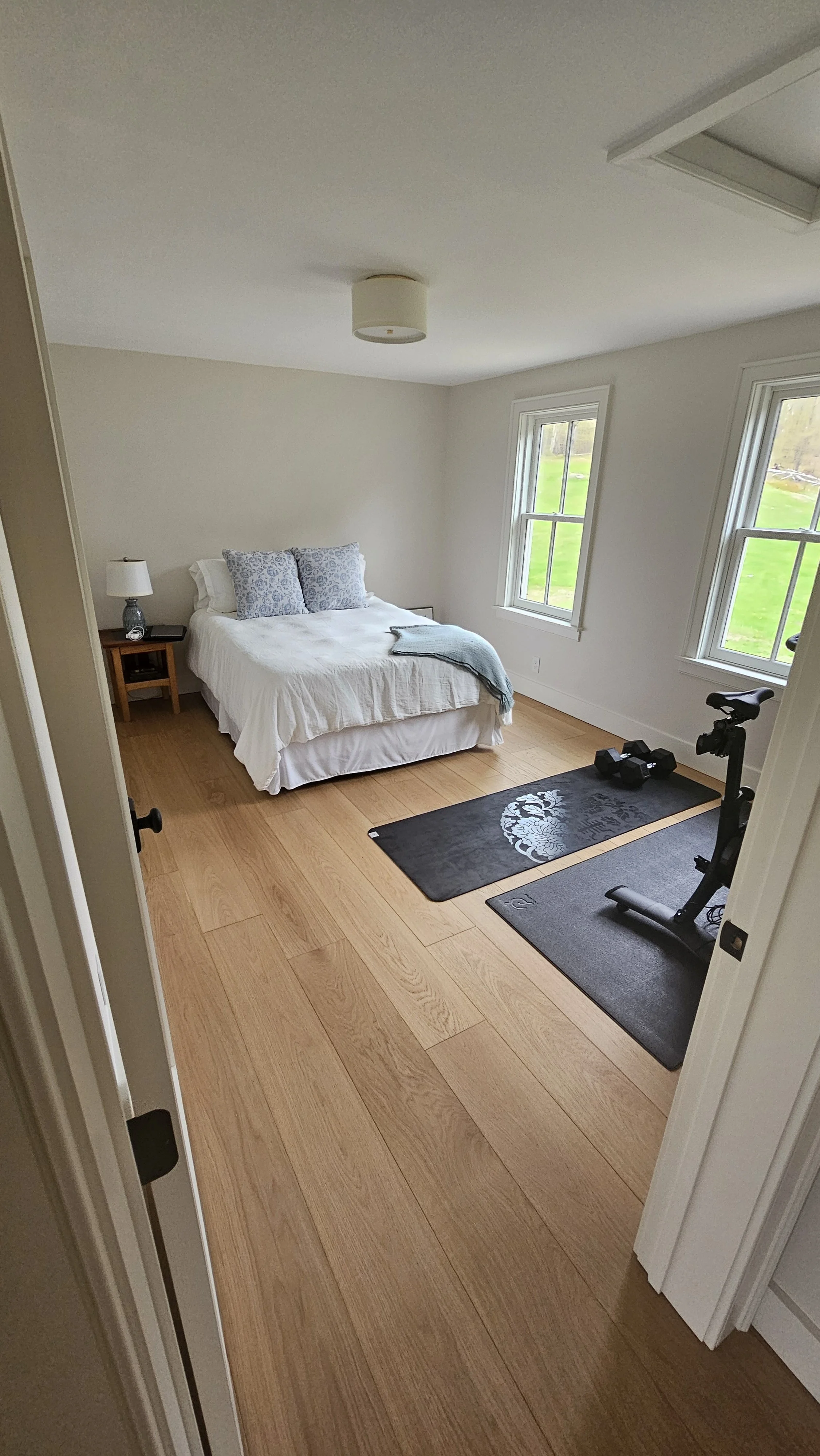 A bedroom with a white bed, two blue patterned pillows, a nightstand with a lamp, two windows showing green grass outside, a black yoga mat with a damask pattern, a folded blanket, and a stationary exercise bike.