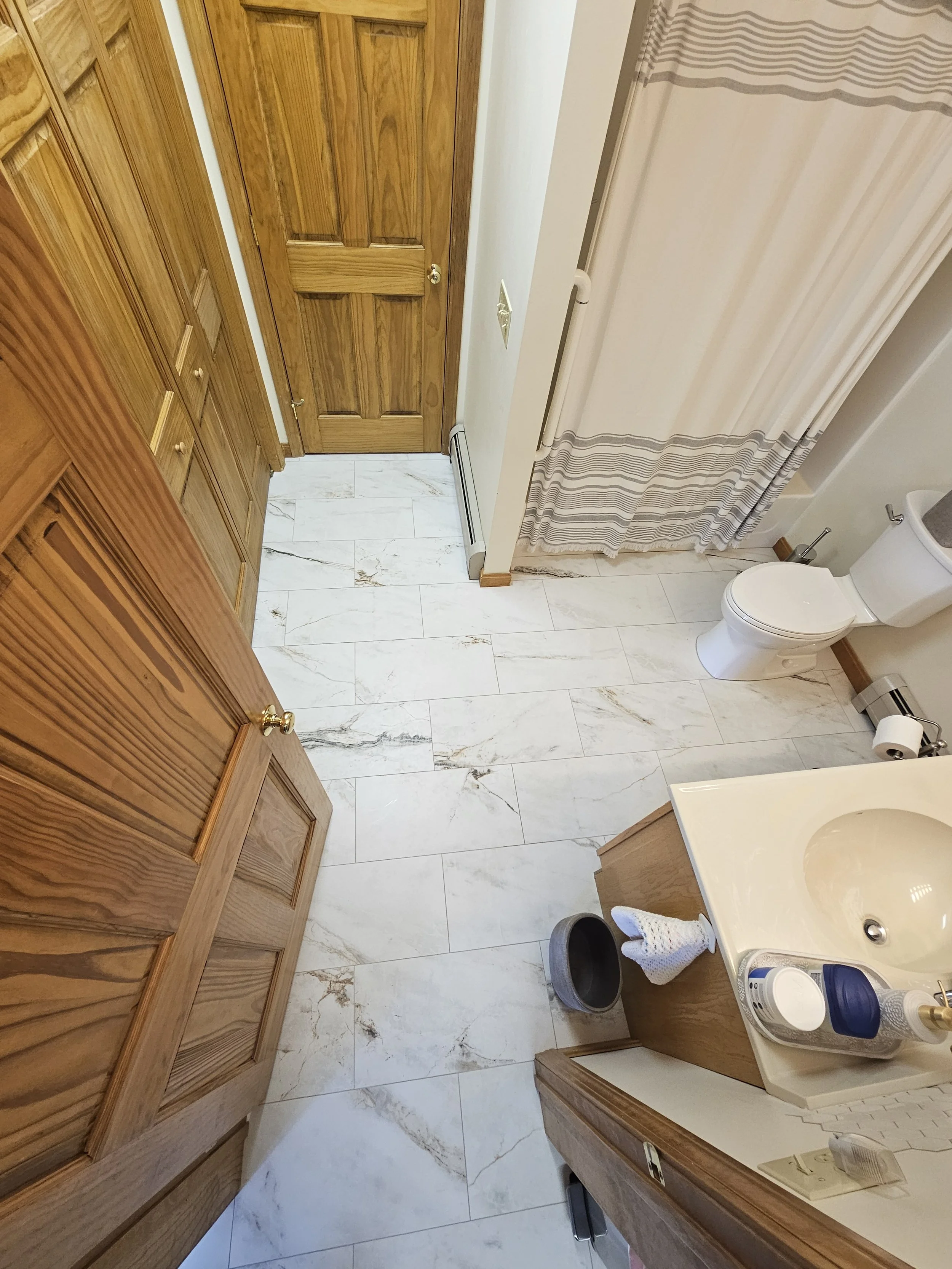 A small bathroom with a wooden door, wooden cabinets, white marble floor, a toilet, a shower curtain, a small sink, and various toiletries and towels.