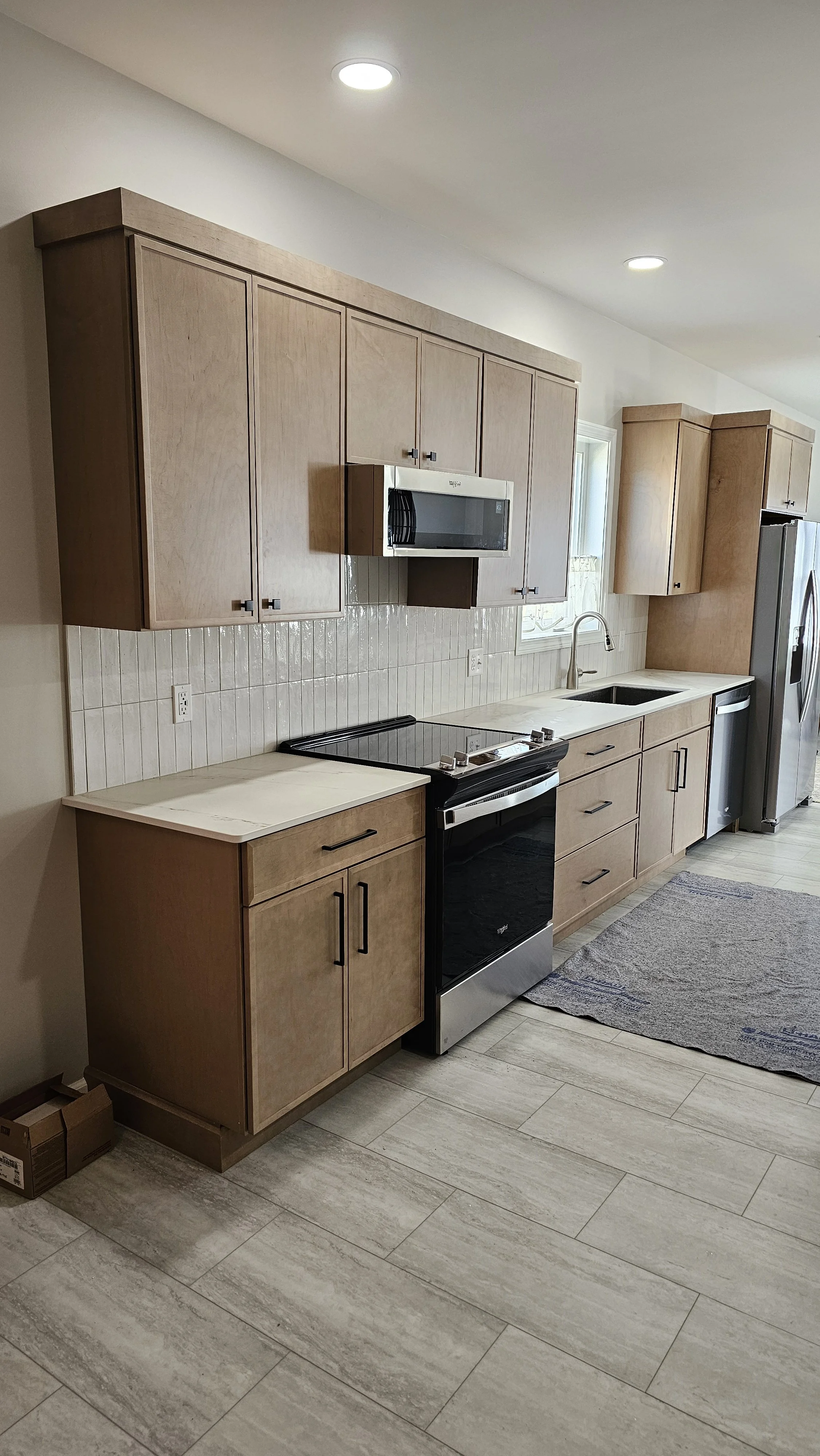A modern kitchen with beige wooden cabinets, a black stove, a silver microwave, a double sink, and a window with natural light, with a gray patterned rug on the floor.