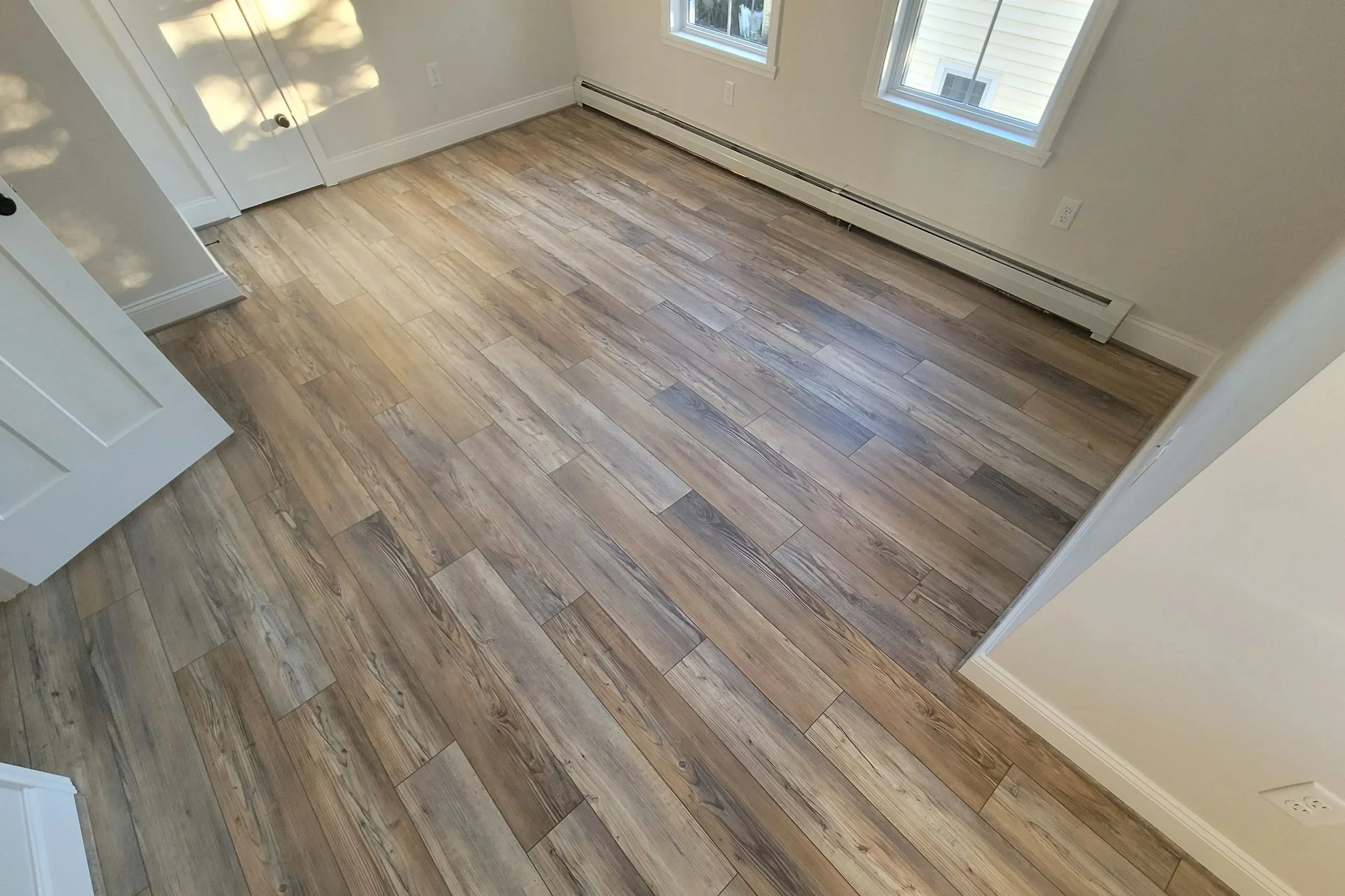 Empty room with light wood flooring, two windows, and a baseboard heater, with a door partially visible.