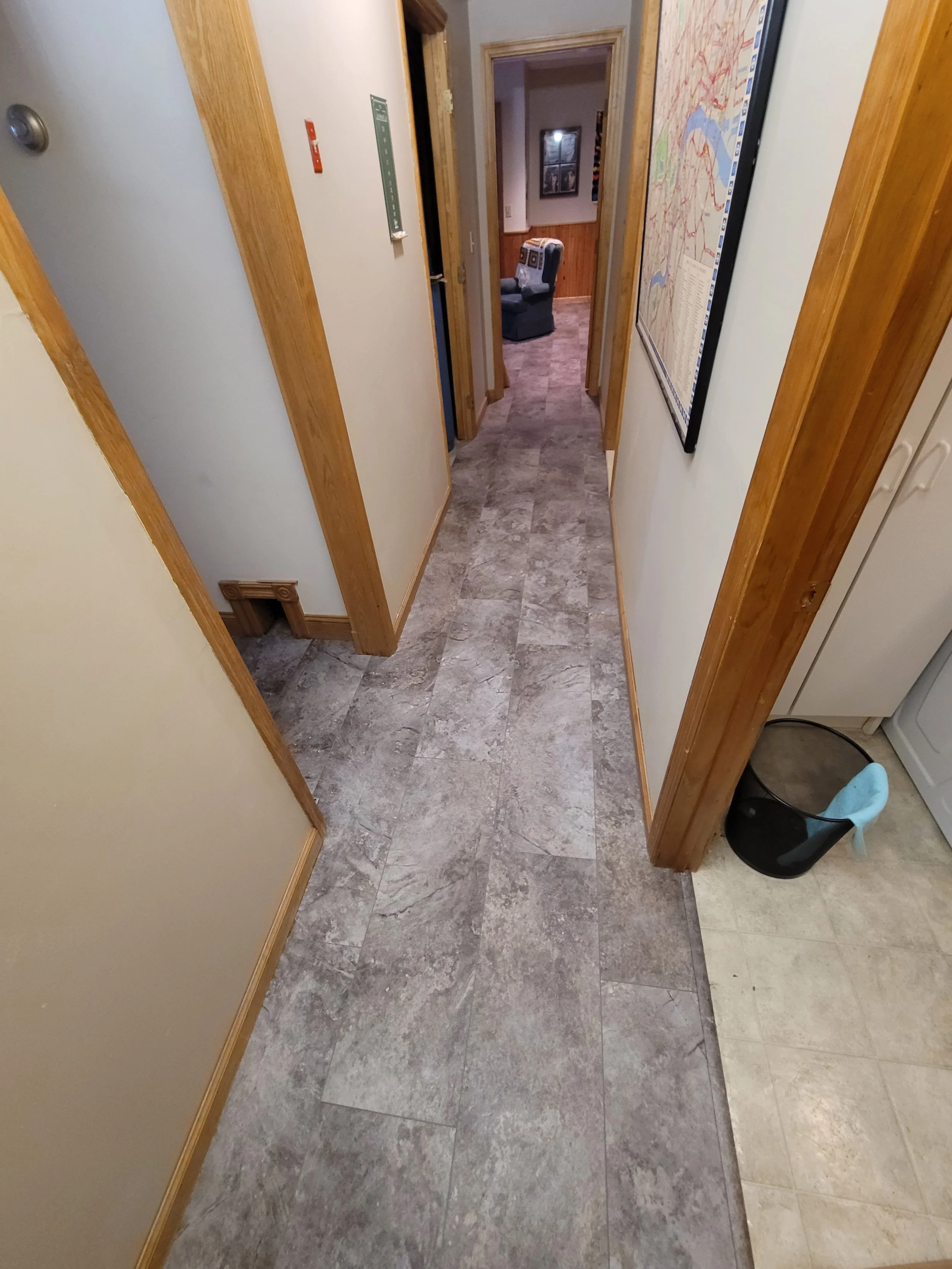 A hallway with a tiled floor, wood trim, and a view into a cozy room with microwaves and framed pictures on the wall.