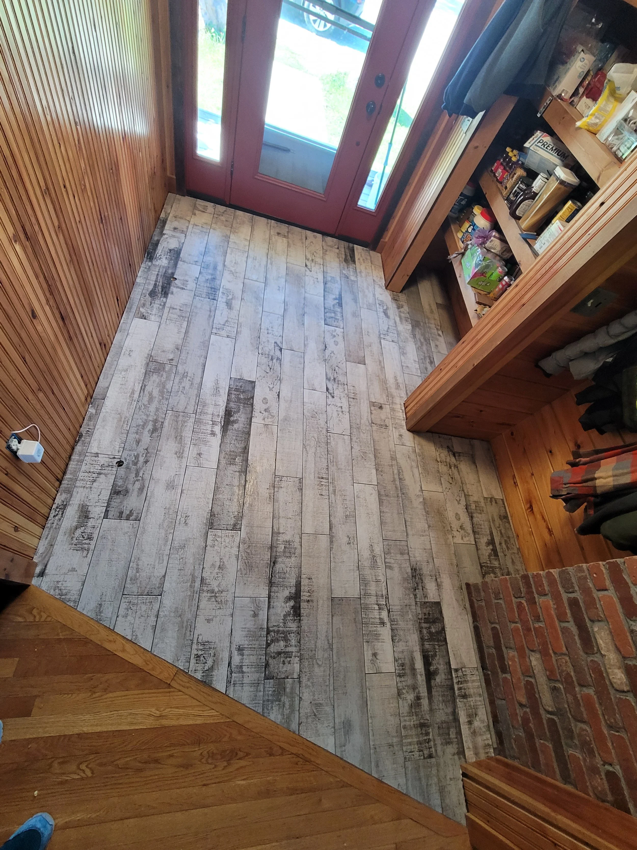 Small entryway with wood-paneled walls, a brick fireplace on the right, a wooden shelf filled with various items, and a wooden floor transitioning to light wood flooring near a pink door with glass panels leading outside.