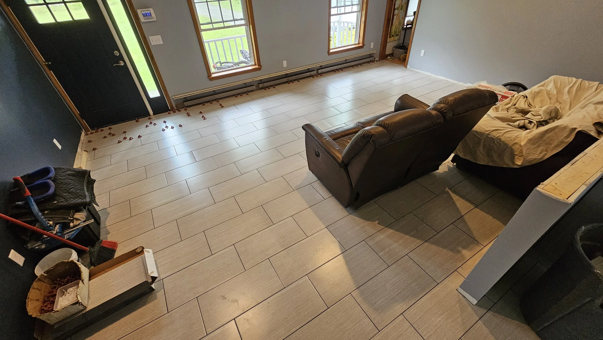 Living room with tan tile flooring, gray walls, and three large windows. A black door is visible on the left, with a small balcony outside one window. There is a brown leather couch and a covered chair in the room, with some miscellaneous items and t