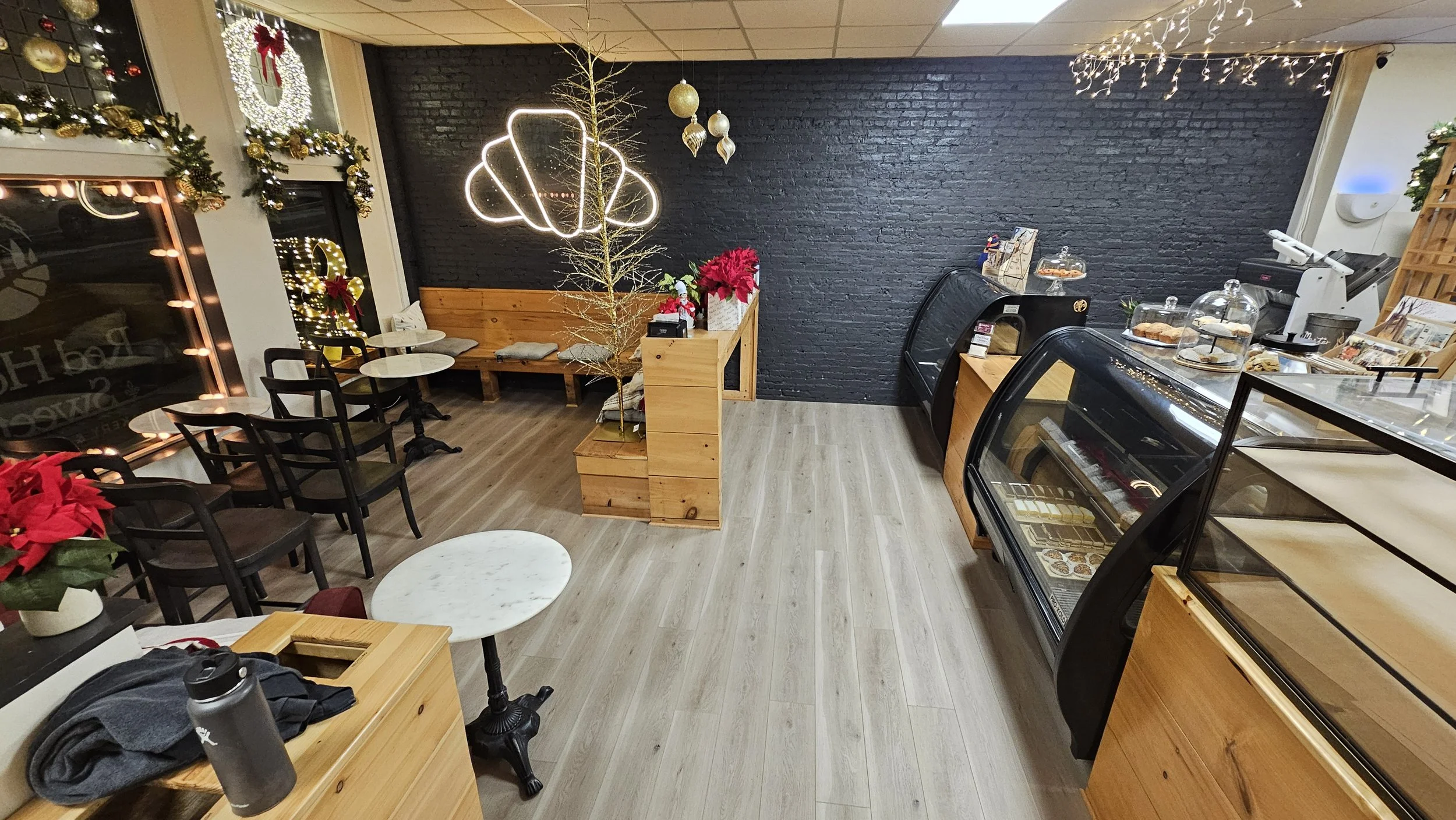 Interior of a cozy cafe decorated with Christmas ornaments and lights, featuring a black brick wall, wooden furniture, a small tree, and display cases with baked goods.