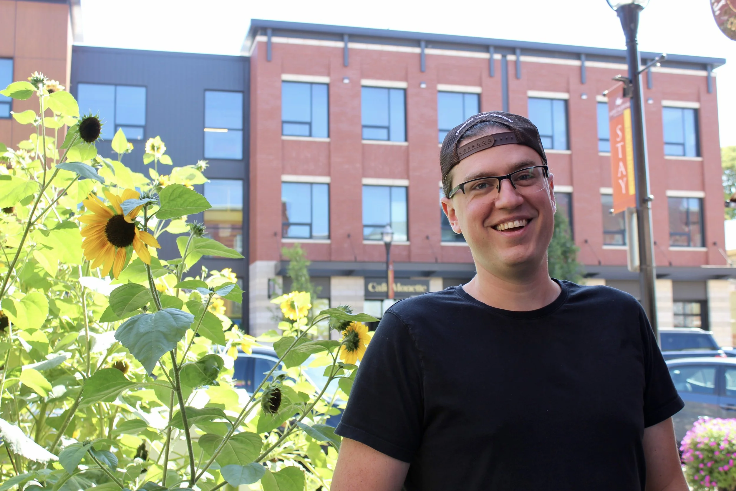 A young man with glasses wearing a backward baseball cap and a black t-shirt standing outdoors in front of a building with a sign that says 'Calligraphy'. Bright yellow sunflower flowers and green foliage are in the foreground, partially obscuring the man.