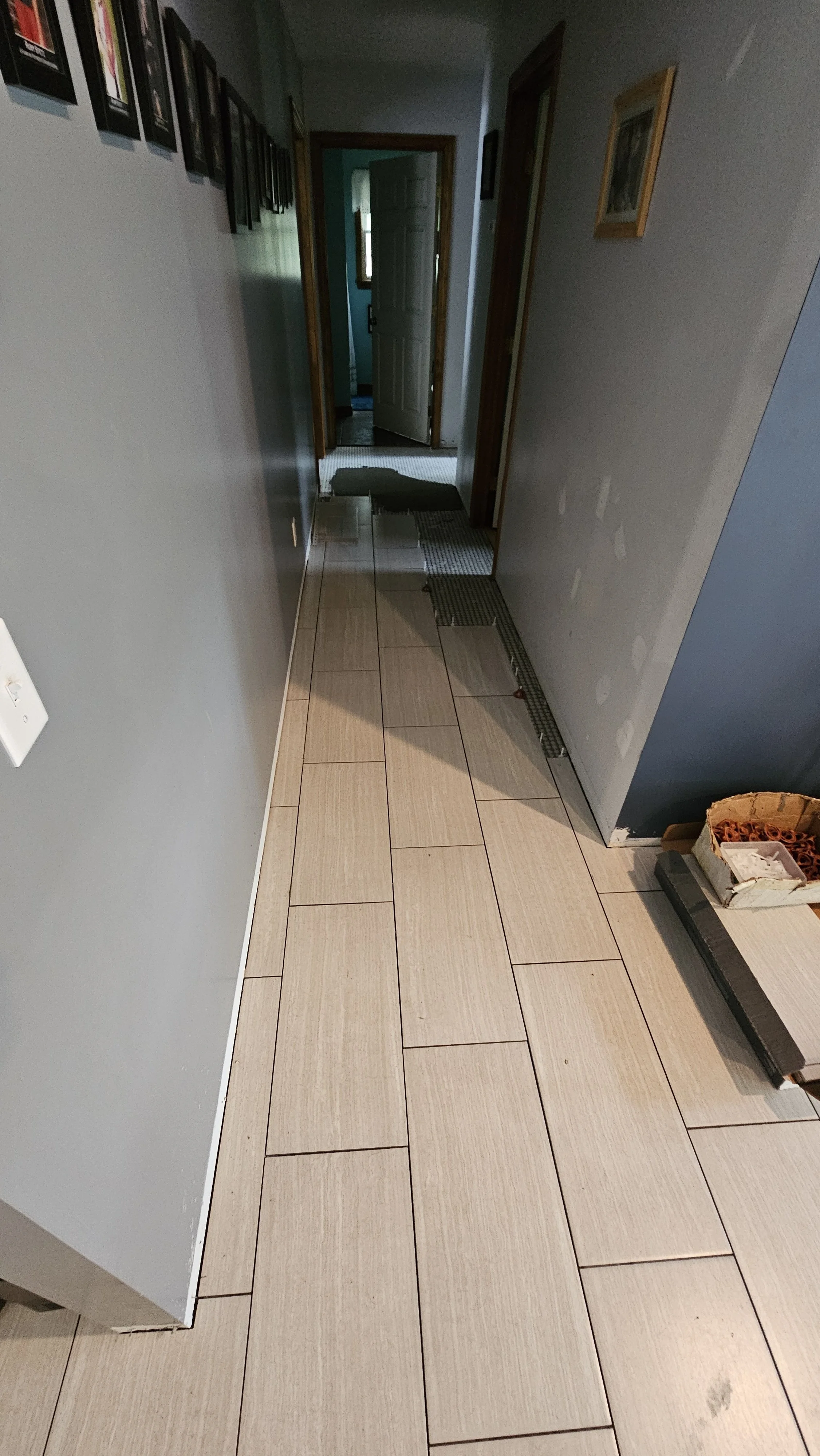 Hallway with beige tiled floor, gray wall on the left, blue wall on the right, and a partially open door at the end revealing another room with teal walls and a window.