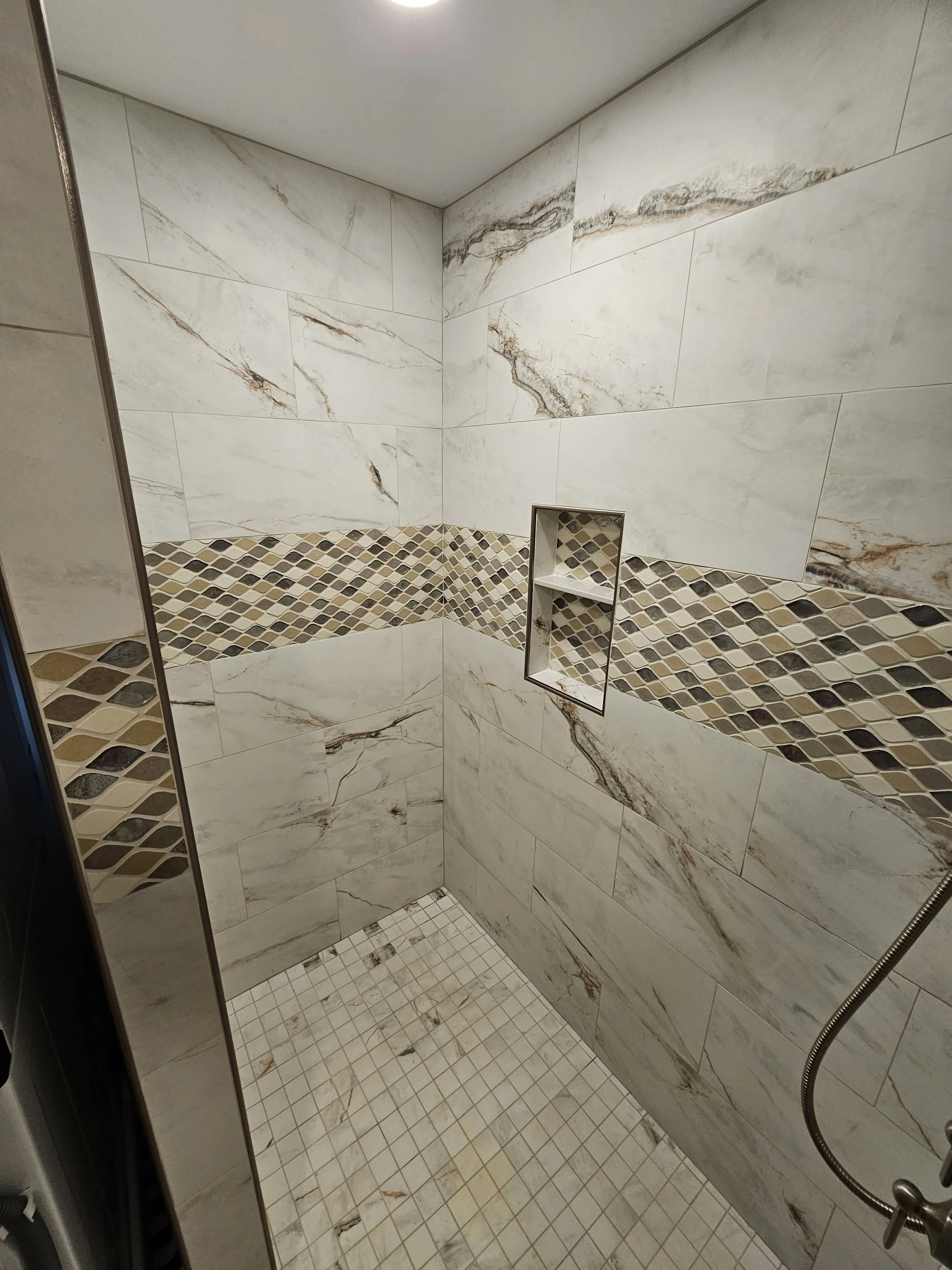 A walk-in shower with large marble-patterned wall tiles, a small tiled floor, and a built-in shelf in the corner.