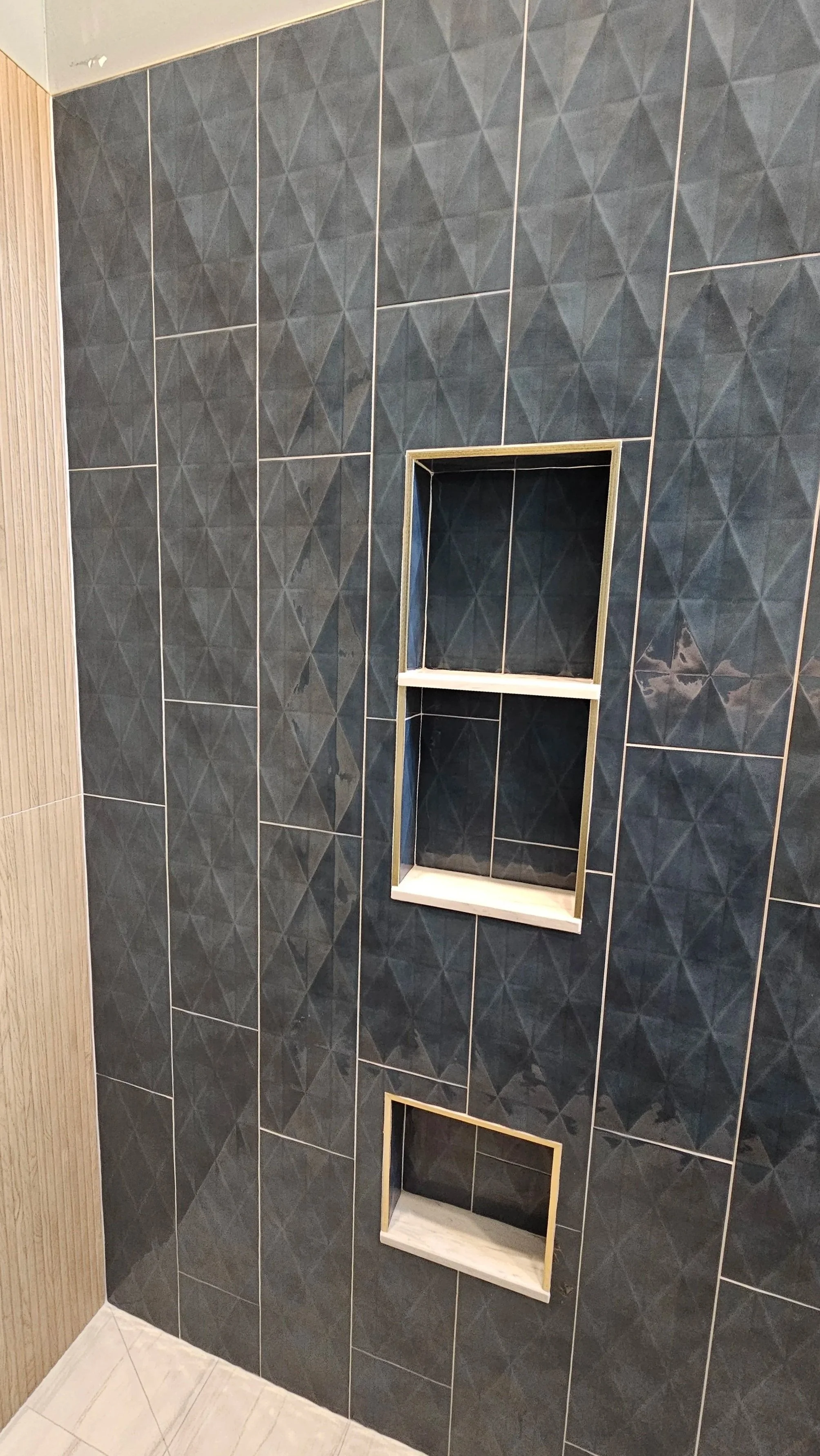 A bathroom wall with dark gray geometric patterned tiles and two built-in shelves framed with light-colored wood.