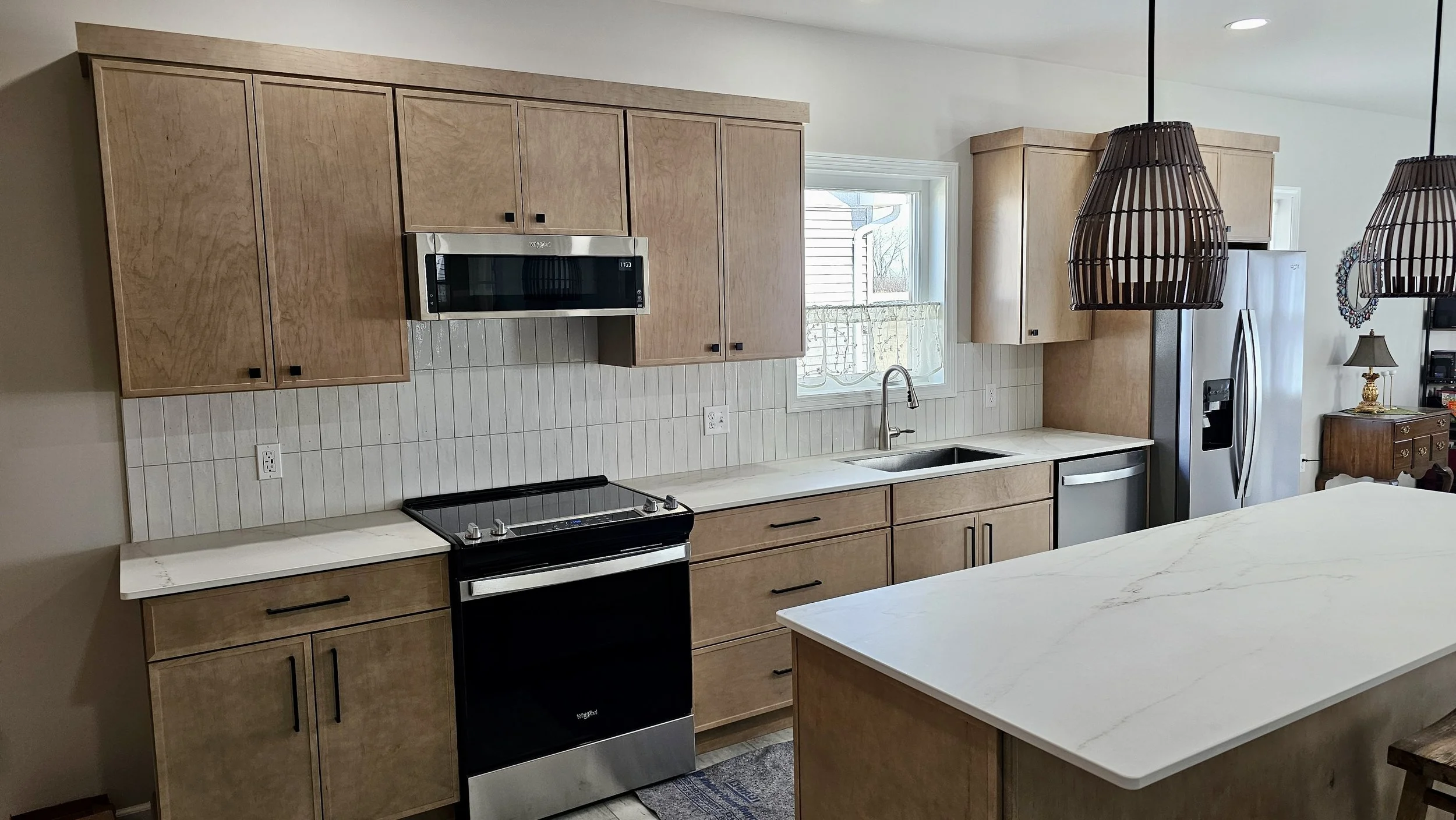 Kitchen with light wood cabinets, white countertops, a window, and appliances including a microwave, stove, dishwasher, and refrigerator, with two hanging pendant lights overhead.