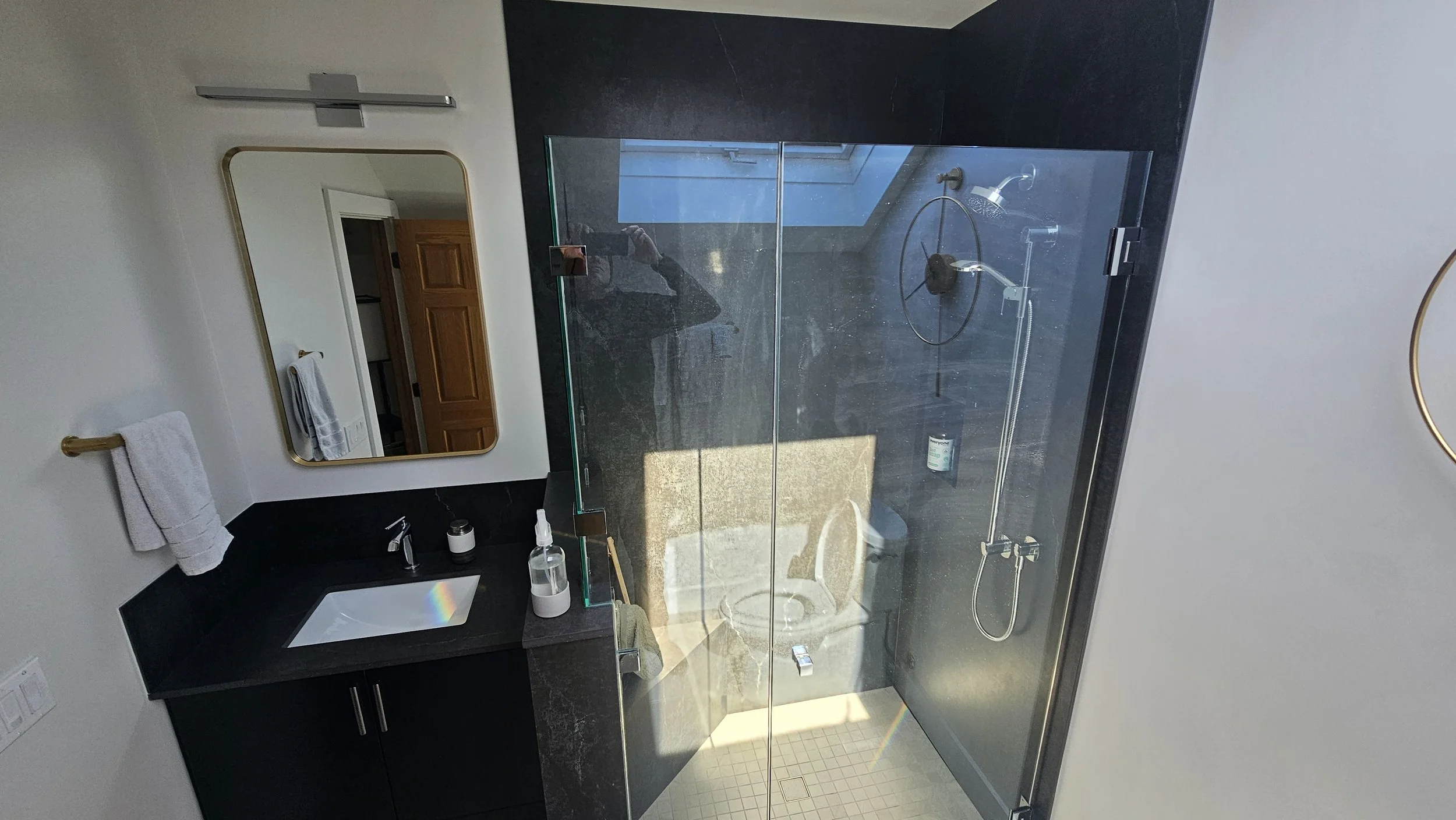Modern bathroom with black and white decor, a glass-enclosed shower with a rain shower head, white towels on a gold towel bar, a mirror, and a black vanity with a white sink, personal care items on the counter.