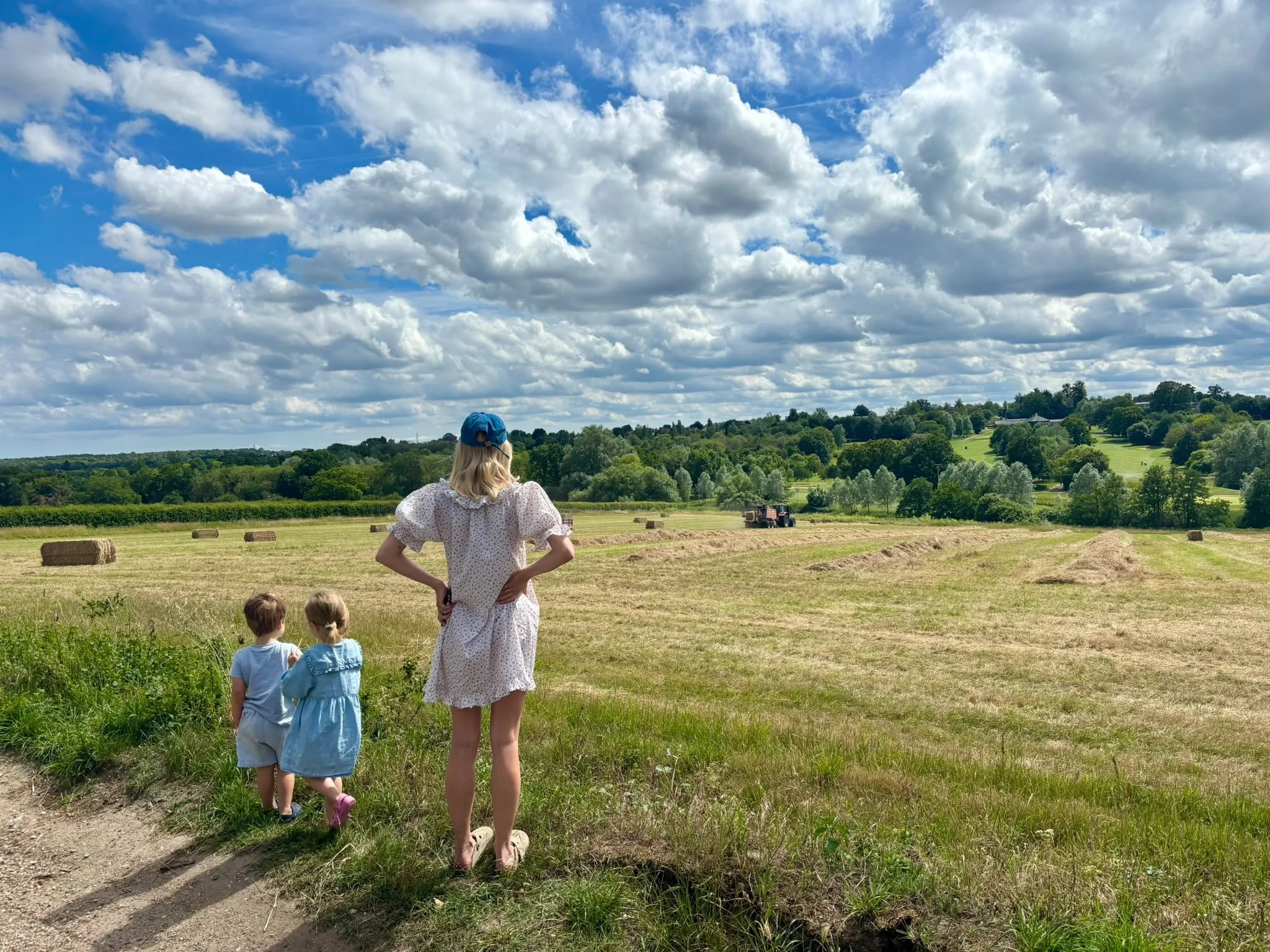 A woman with blonde hair in a floral dress and blue cap is standing in a field with children, looking out at a lush green landscape under a partly cloudy sky with clouds and blue patches.