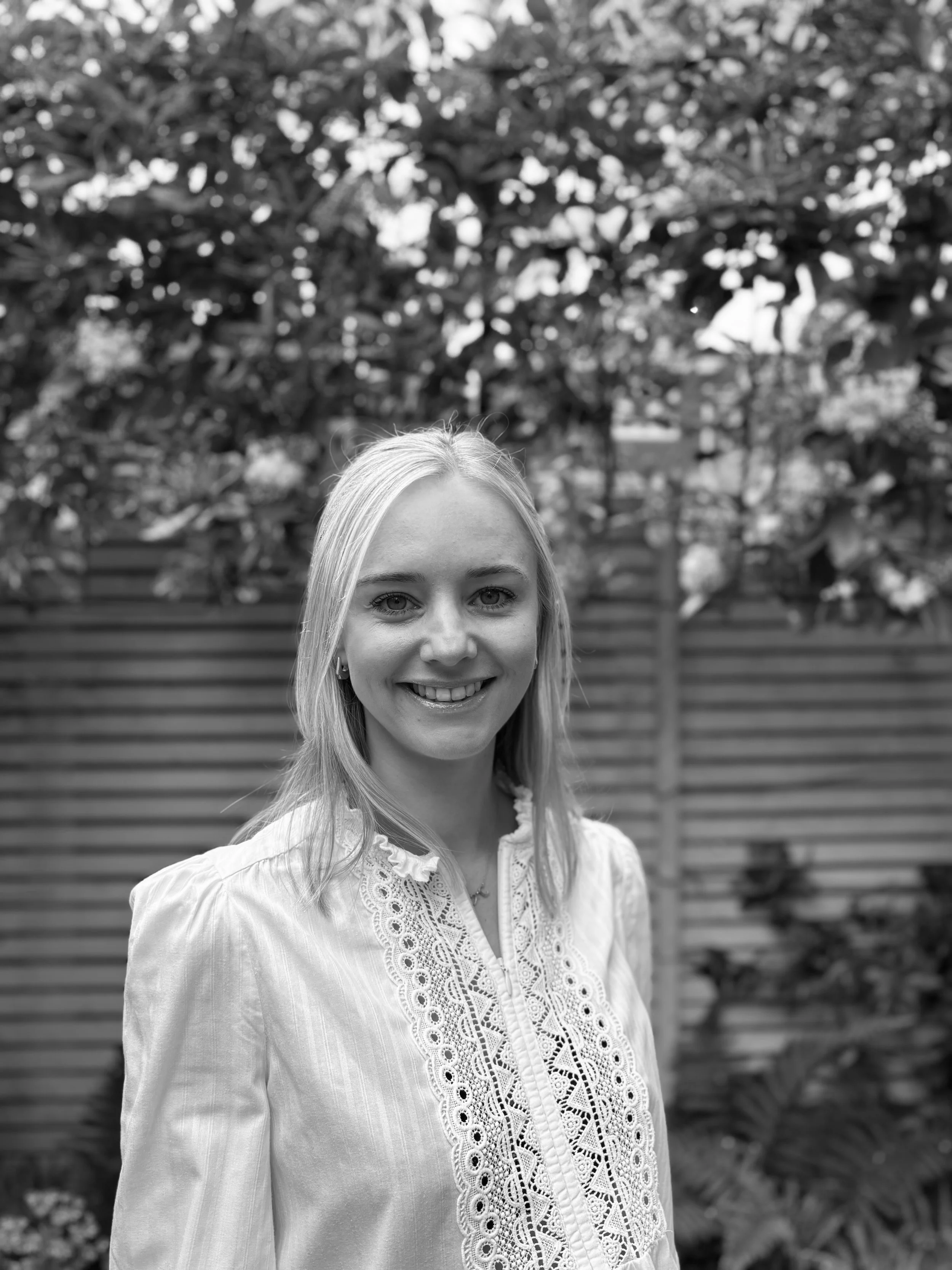 A woman with long light hair smiling outdoors in front of a tree and wooden fence, wearing a white embroidered blouse.