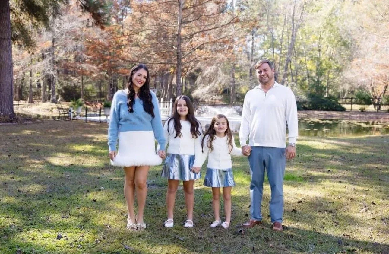 A family of five holding hands and posing outdoors by a pond in a park with trees and autumn foliage.
