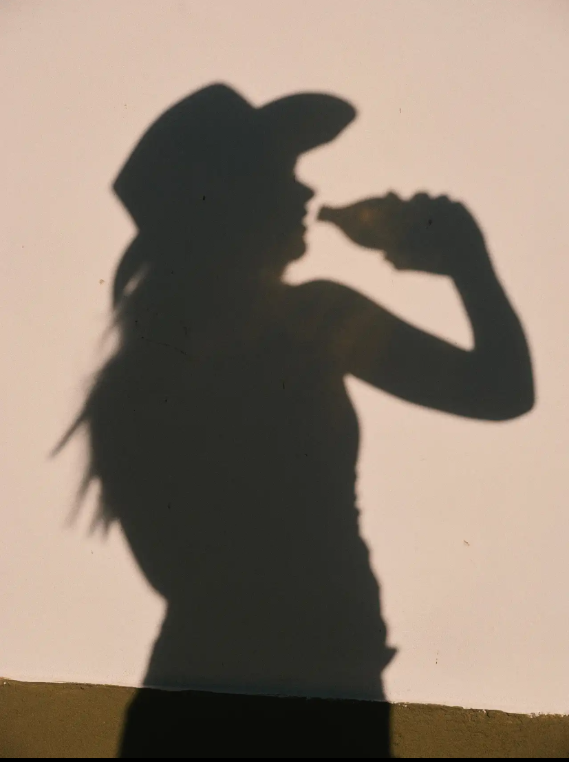 Silhouette of a woman wearing a cap, holding a snack near her mouth, with her hair visible and a plain background.