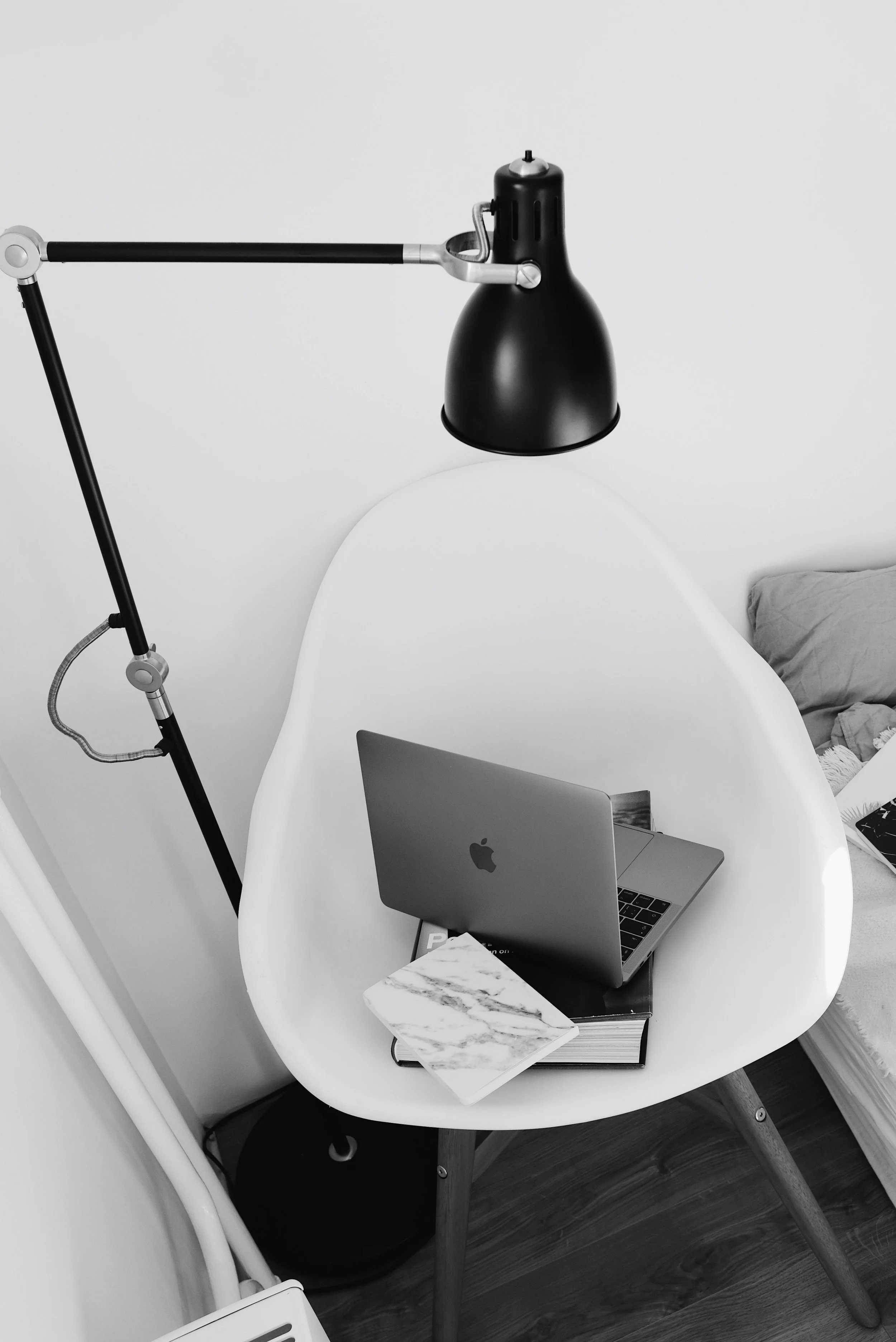 Mesa blanca con una laptop de marca Apple, un libro con cubierta de mármol y una libreta debajo del libro. En la esquina de la mesa hay una libreta más pequeña con cubierta blanca y negra. Al fondo, hay una cama con almohada y una luz de escritorio negra con brazo articulado en el costado de la mesa.