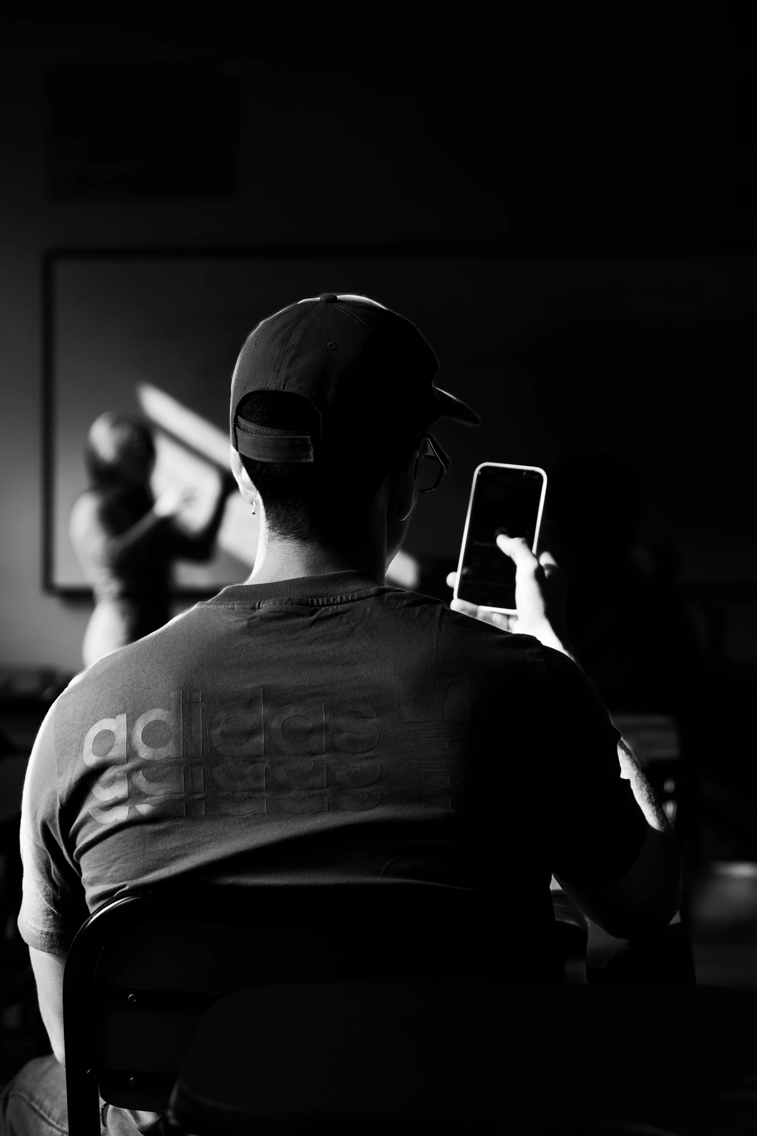 Una persona con gorra toma una selfie en un espejo, mientras otra persona en el fondo parece ajustar la iluminación o el maquillaje.
