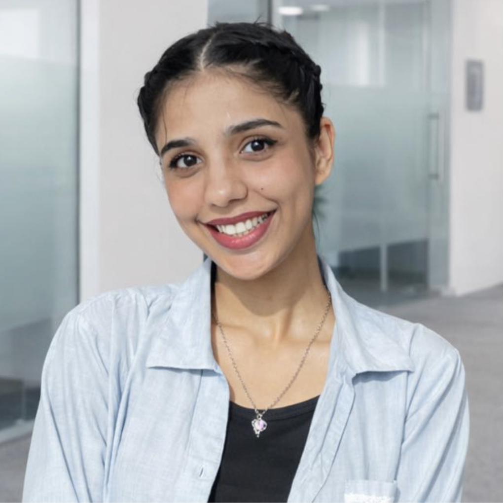 A young woman with braided dark hair smiling, wearing a light-colored shirt over a black top and a necklace, in a modern office environment.