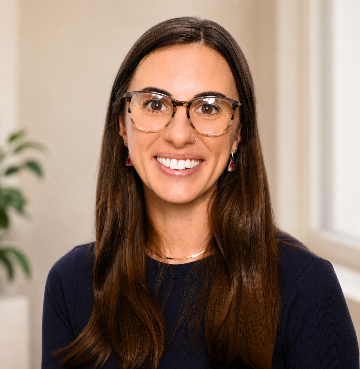 A woman with long brown hair wearing glasses smiling at the camera in a well-lit room.