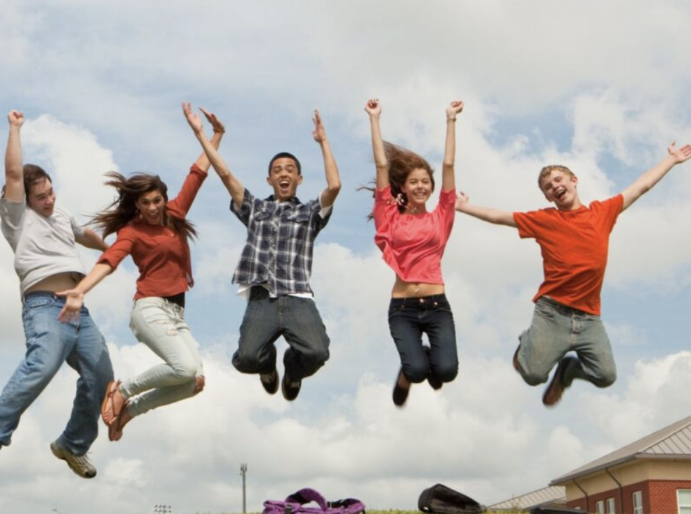 Five young people jumping in the air outdoors on a partly cloudy day, smiling and having fun.