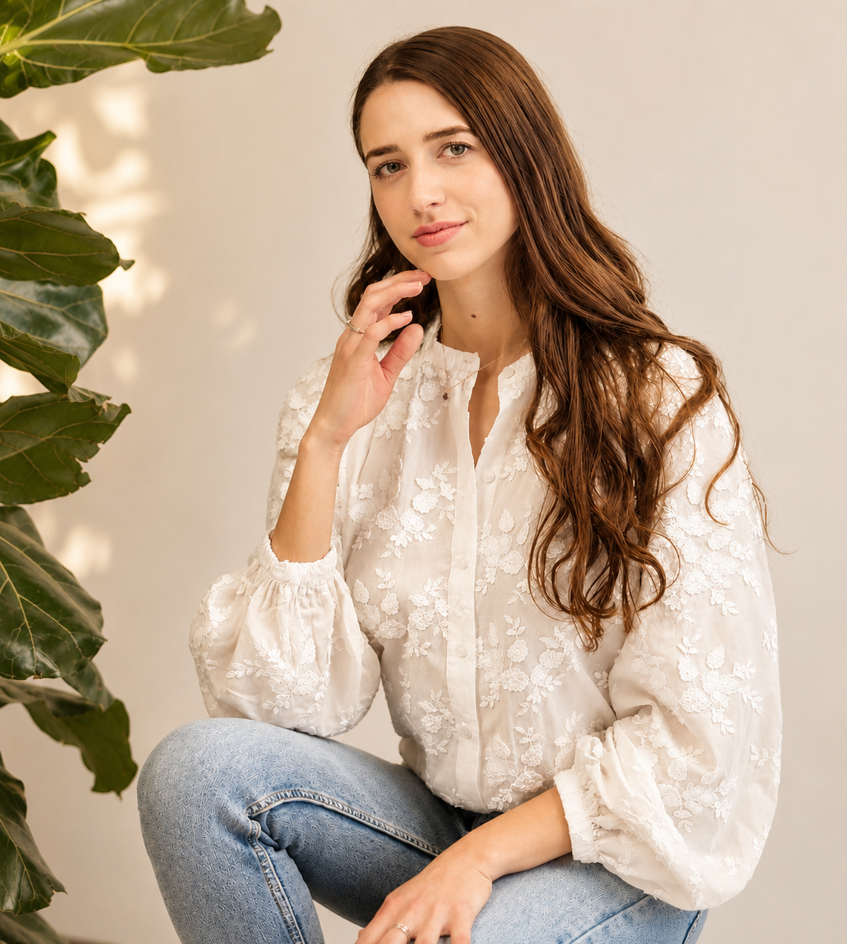 A young woman with long wavy brunette hair, sitting near a large green plant, wearing a white embroidered blouse and light blue jeans, looking at the camera with a gentle smile.
