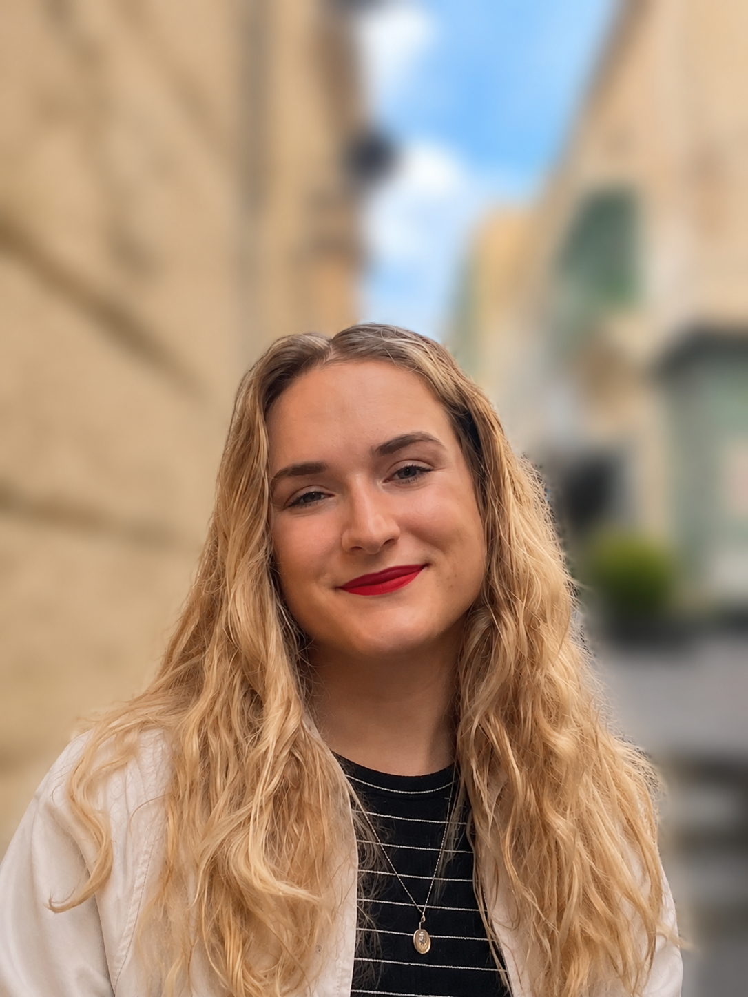 A young woman with long curly blonde hair, wearing red lipstick, a black striped shirt with a necklace, and a cream jacket, standing outdoors with a blurred beige building and blue sky in the background.