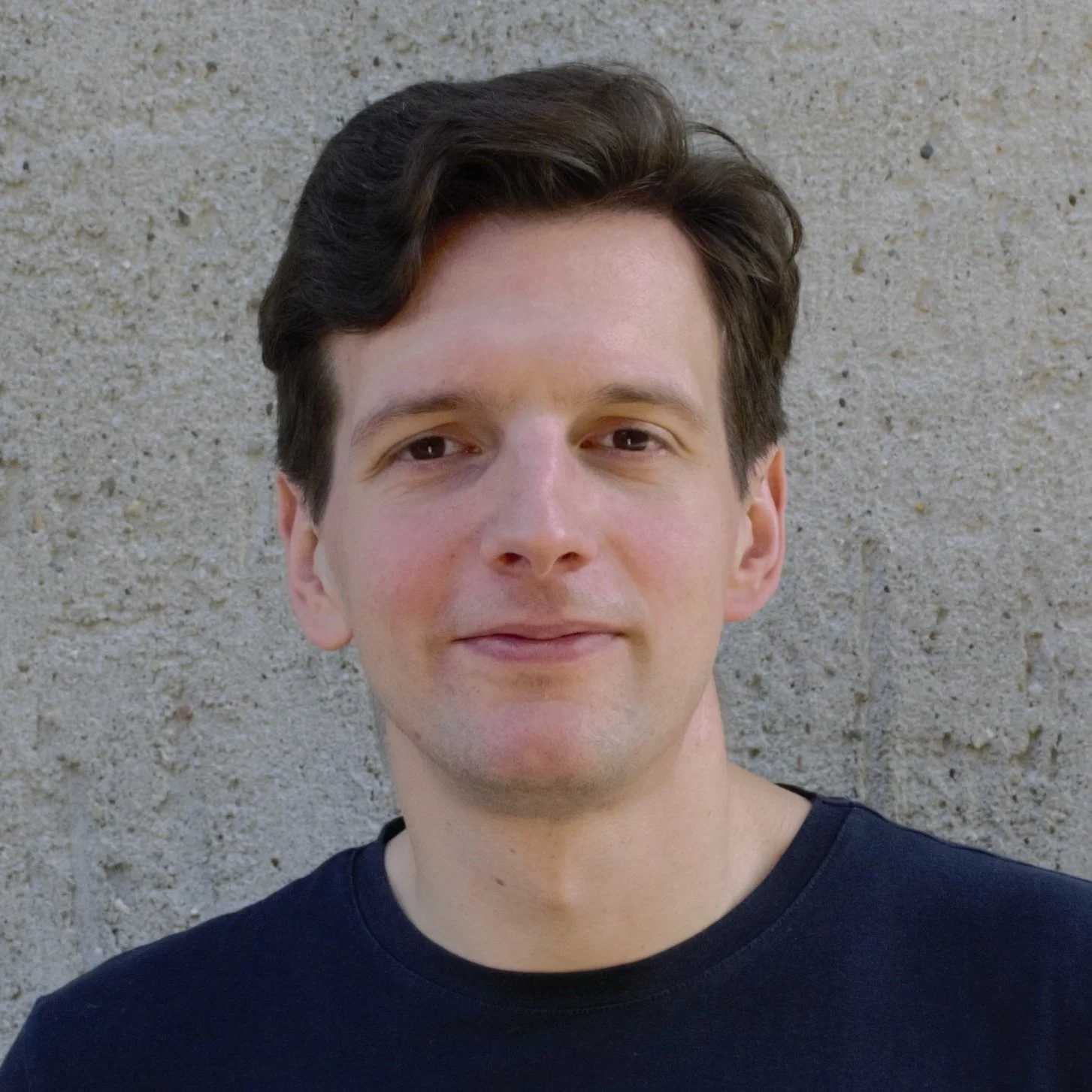 A young man with dark brown hair, fair skin, and a slight smile, wearing a black shirt, standing against a beige textured wall.