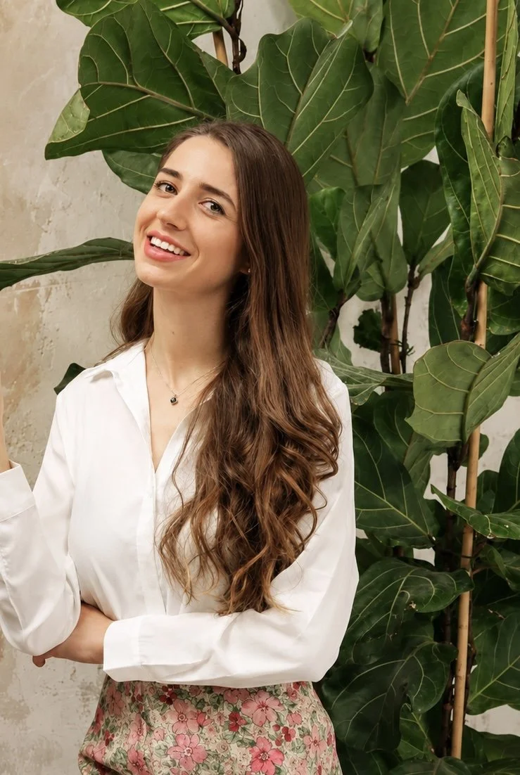 A young woman with long, wavy brown hair smiling, standing in front of large green leaves. She wears a white blouse and a floral skirt.