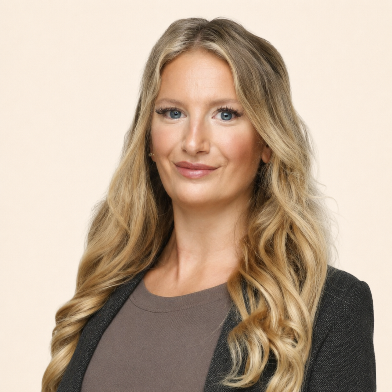 Portrait of a woman with long wavy blonde hair, blue eyes, wearing a black blazer over a gray top, smiling softly against a plain light background.