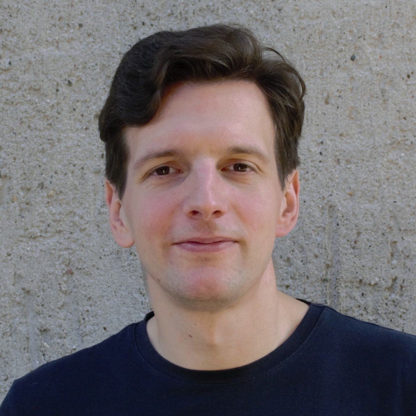 A young man with dark hair and fair skin standing outdoors against a beige concrete wall, wearing a dark t-shirt, smiling slightly at the camera.