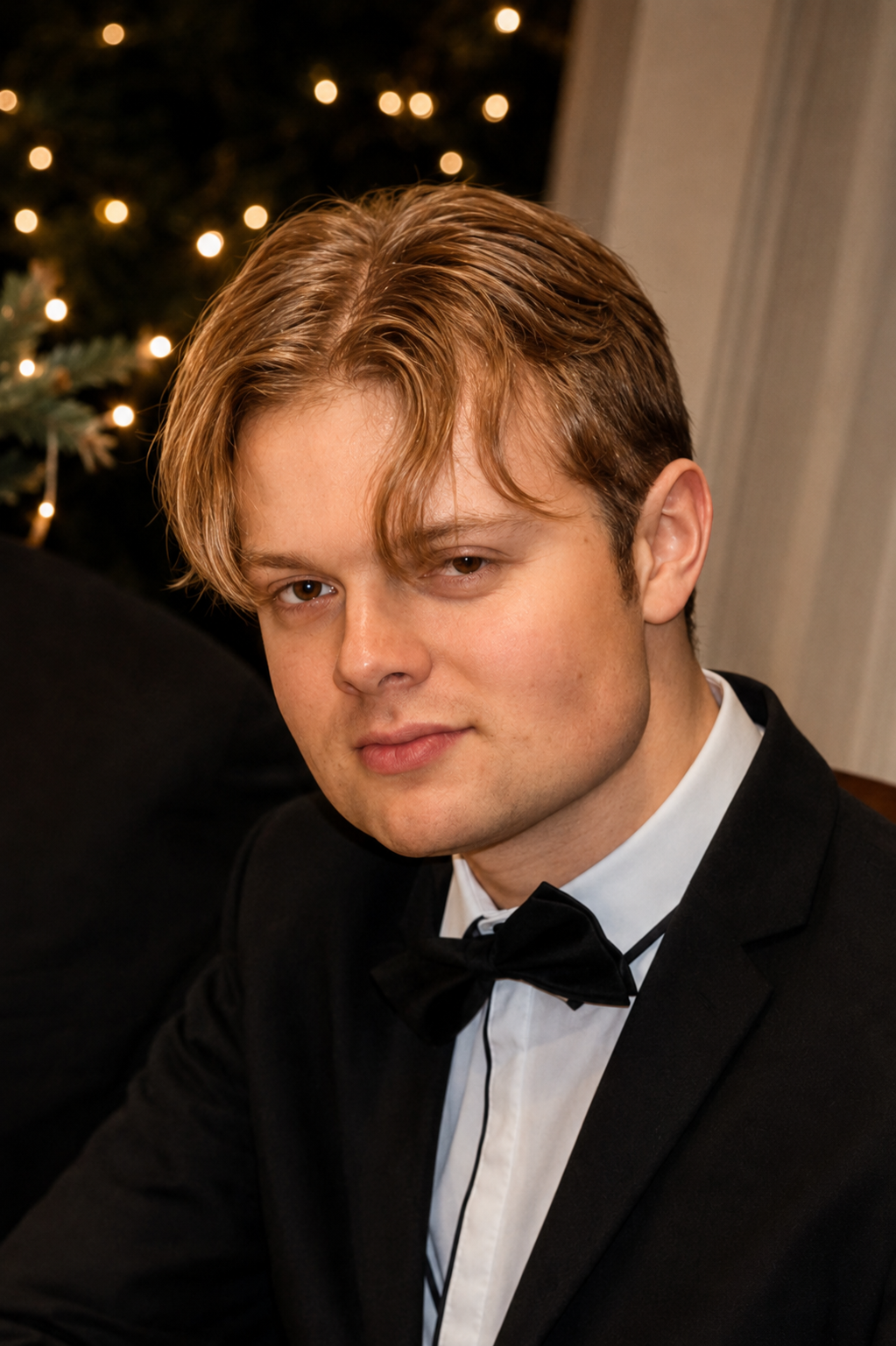 A young man with light brown hair in a tuxedo and bowtie, sitting indoors near a decorated Christmas tree with lights.