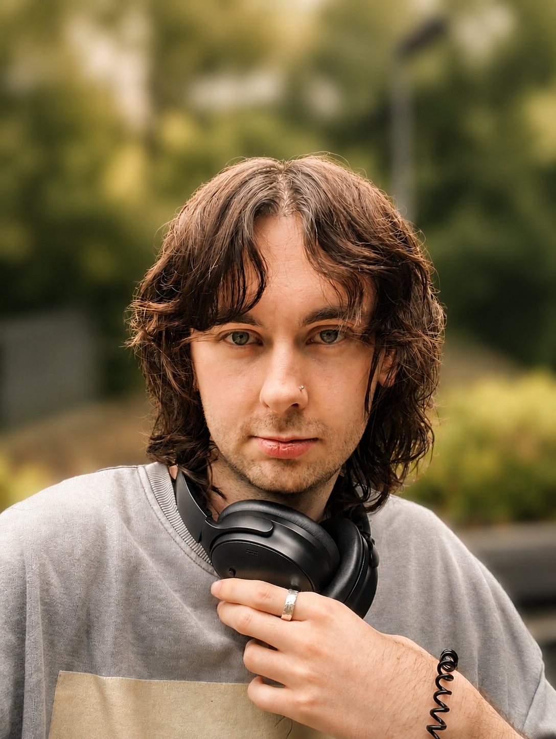 A young man with wavy brown hair, green eyes, and light skin, holding a pair of black headphones around his neck outdoors.