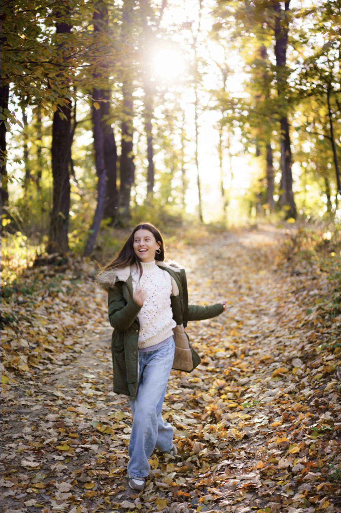 A young woman walking along a leaf-covered trail in a forest during autumn, smiling and enjoying the sunny day.