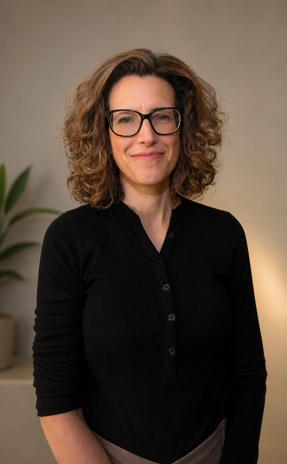 A woman with curly brown hair, wearing black glasses and a black button-up shirt, is smiling in an indoor setting.