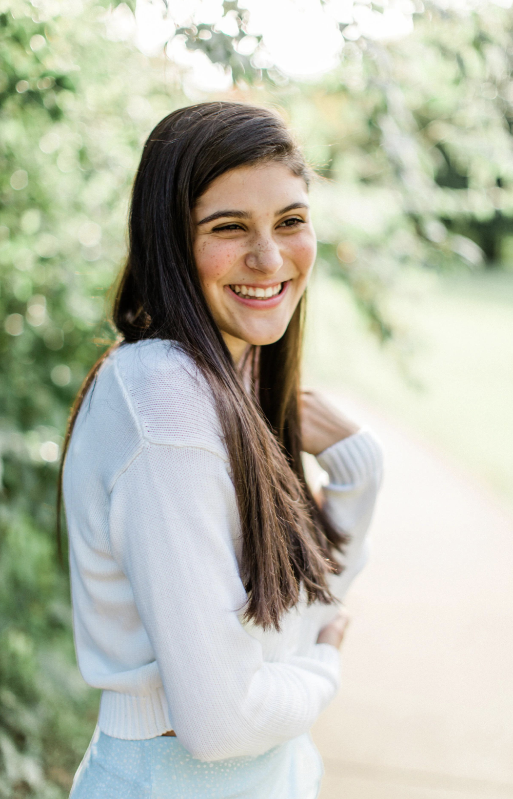 A young woman with long dark hair and freckles, smiling outdoors during daytime with green foliage in the background.