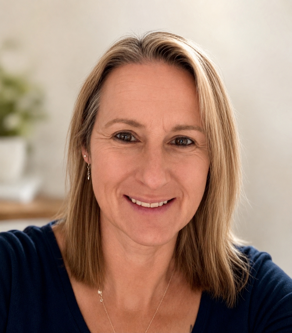 A woman with shoulder-length blonde hair smiling, wearing a dark top and earrings, in a well-lit room with a blurred background of a plant and a light-colored wall.