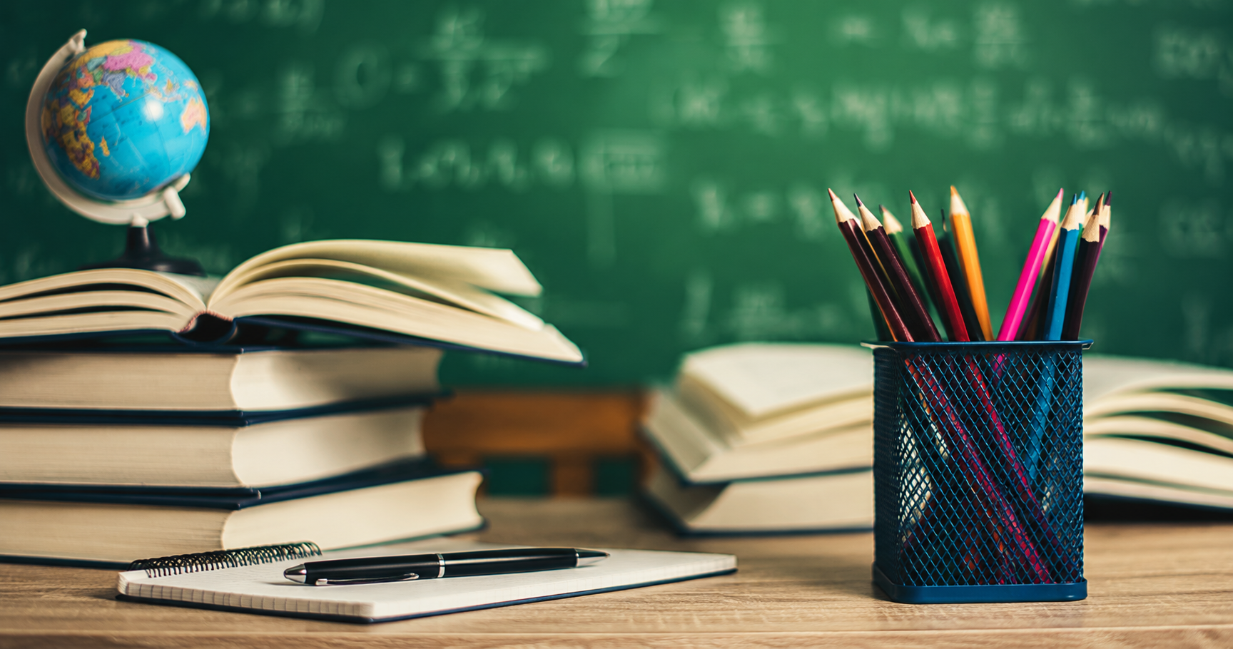 School desk with open books, a small globe, a pen, and a container of colored pencils in front of a chalkboard with writing.
