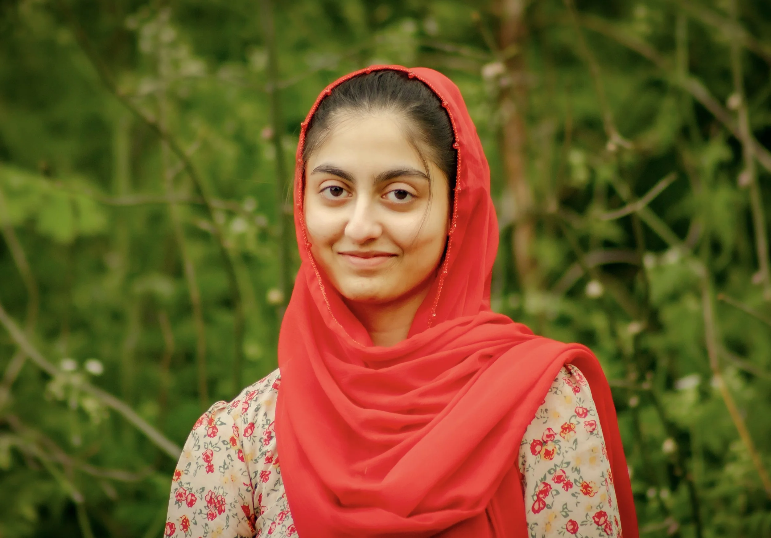 A young woman with dark hair wearing a red headscarf and a floral patterned top stands outdoors with a blurred green foliage background.