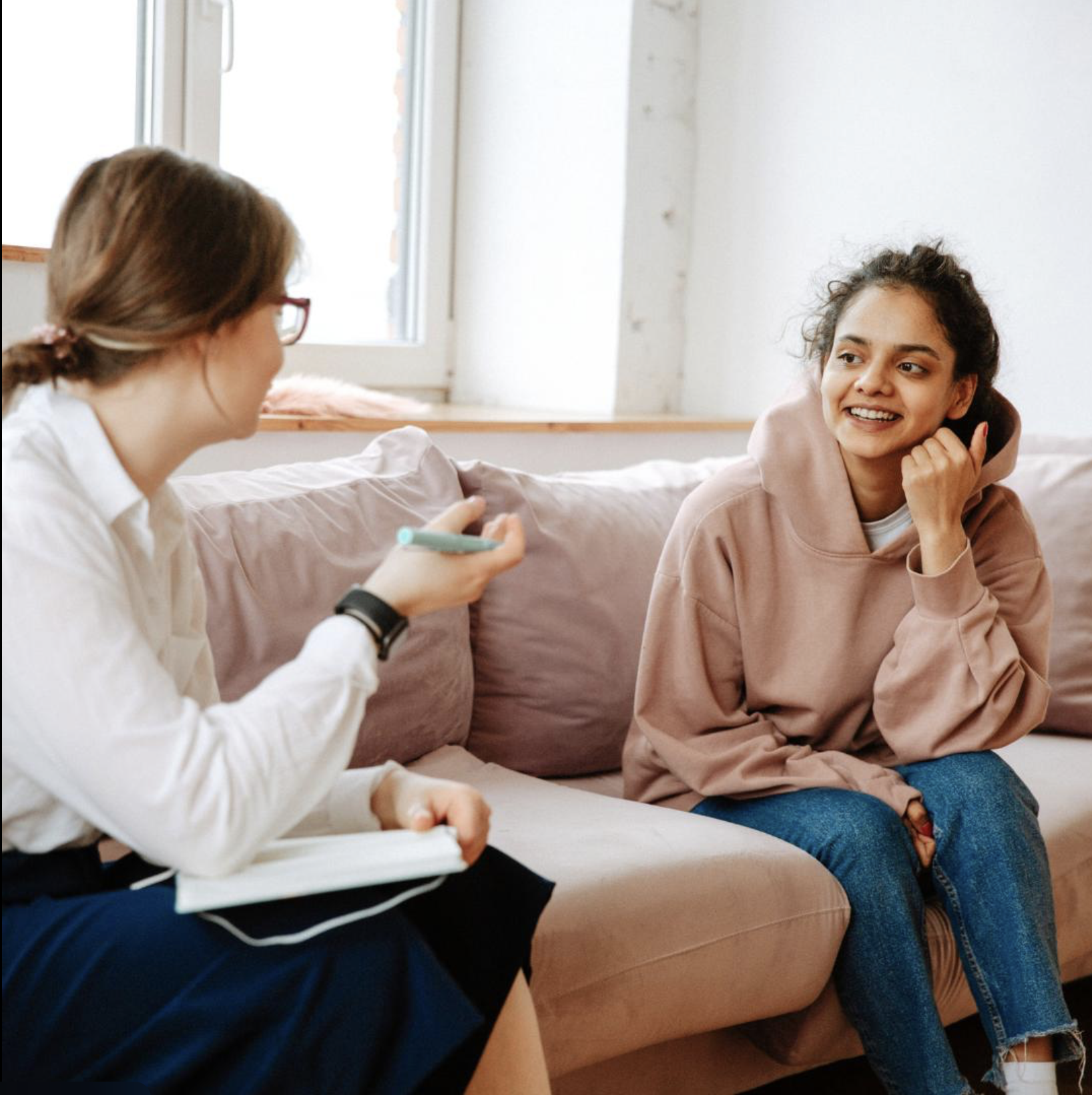 Two women engaged in conversation sitting on a light pink sofa in a bright room with white walls and a window, one wearing a white shirt and glasses, the other in a pink hoodie and blue jeans.