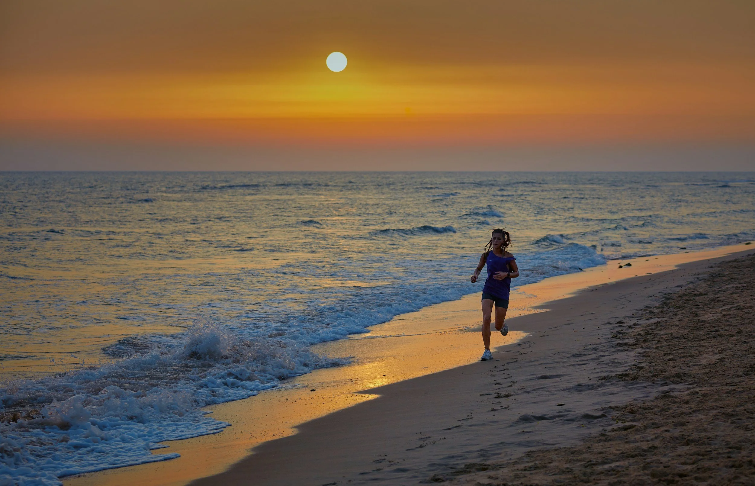 A woman jogging on the beach during sunset, with the sun low on the horizon and the sky painted in orange and purple hues.