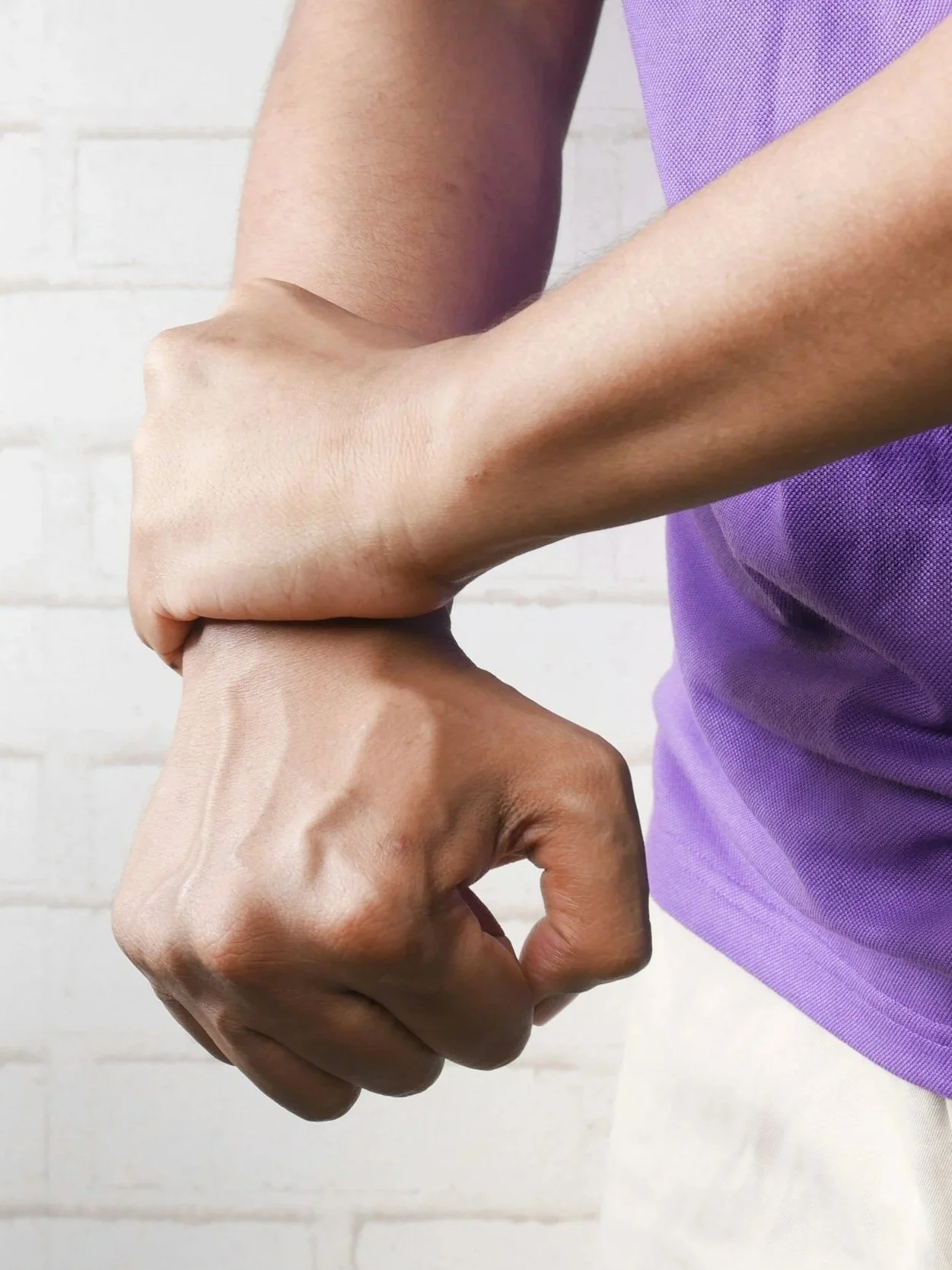 A person with dark skin making a fist and holding their other arm in pain, wearing a purple shirt. The background is a white brick wall.