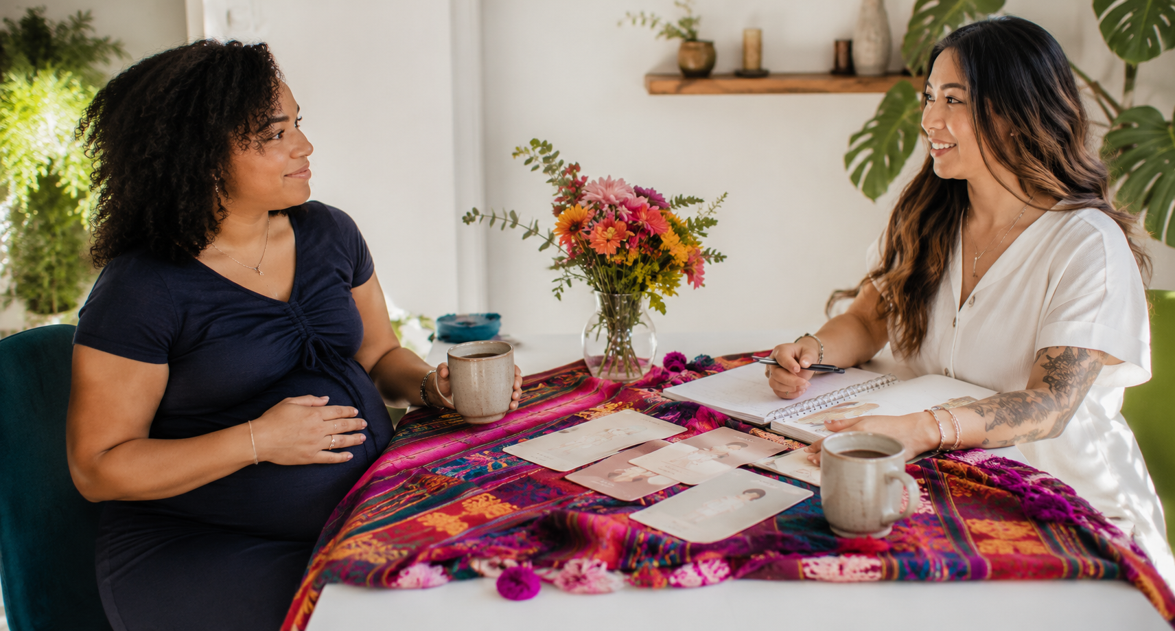 Twee vrouwen zitten aan een tafel met kleurrijke tafelkleed, bloemen in een vaas, papier en pennen, en drinken. Ze praten vriendelijk, een van hen is zwanger.