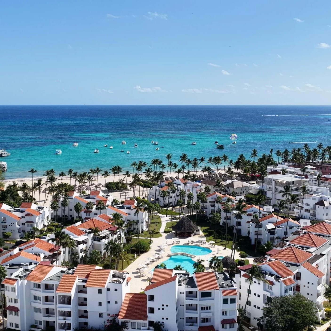 Aerial view of a beachfront resort with white buildings, orange roofs, a swimming pool, lush palm trees, and boats anchored in the turquoise ocean, under a blue sky with scattered clouds.