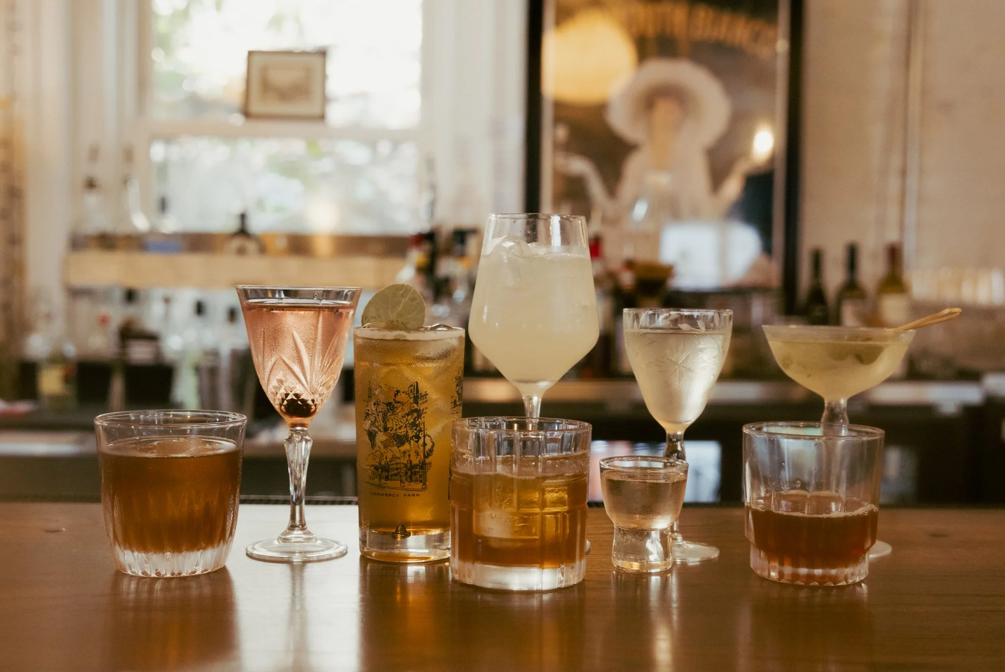 Various cocktails and drinks in different glasses on a wooden table, with a blurred background of a cozy cafe or bar setting.